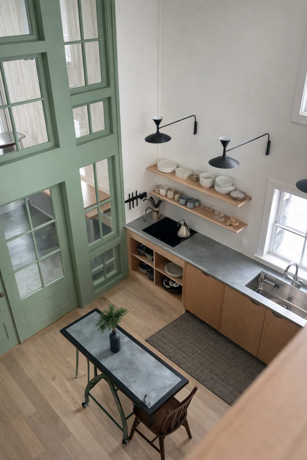 High-angle view of a modern kitchen featuring light wood cabinetry, a gray countertop, stainless steel sink, and an induction cooktop, with tall green-framed windows and doors adding a pop of color.