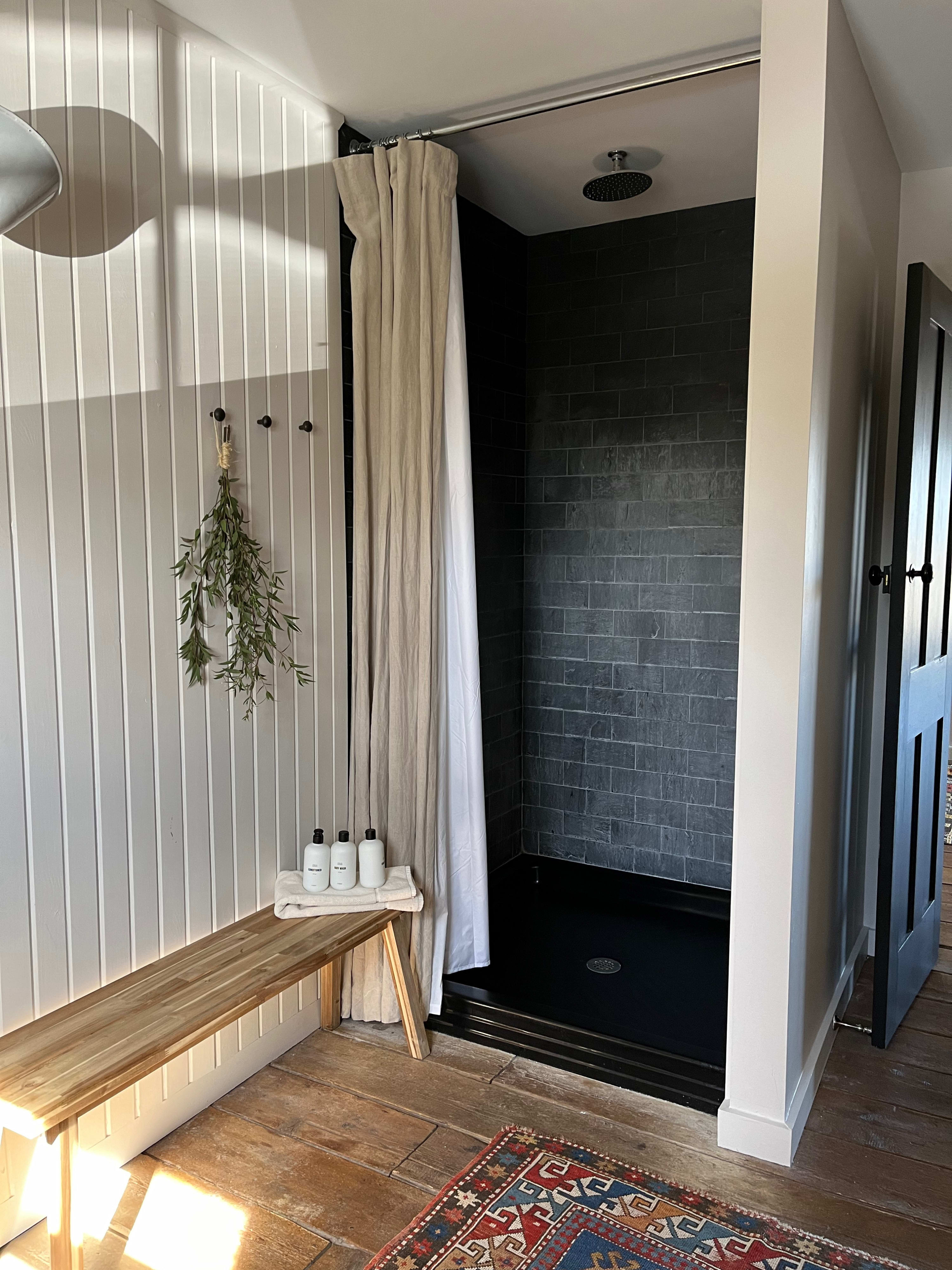A close-up of the walk-in shower in the rustic bathroom, featuring black subway tiles, a dark shower pan, and a light-colored shower curtain. A wooden bench with toiletries sits nearby, against white vertical paneling.