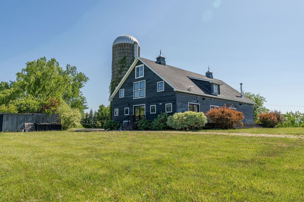 A ground-level view of the dark gray barn-style house with a tall silo, set against a bright blue sky, surrounded by a well-maintained green lawn and various shrubs.