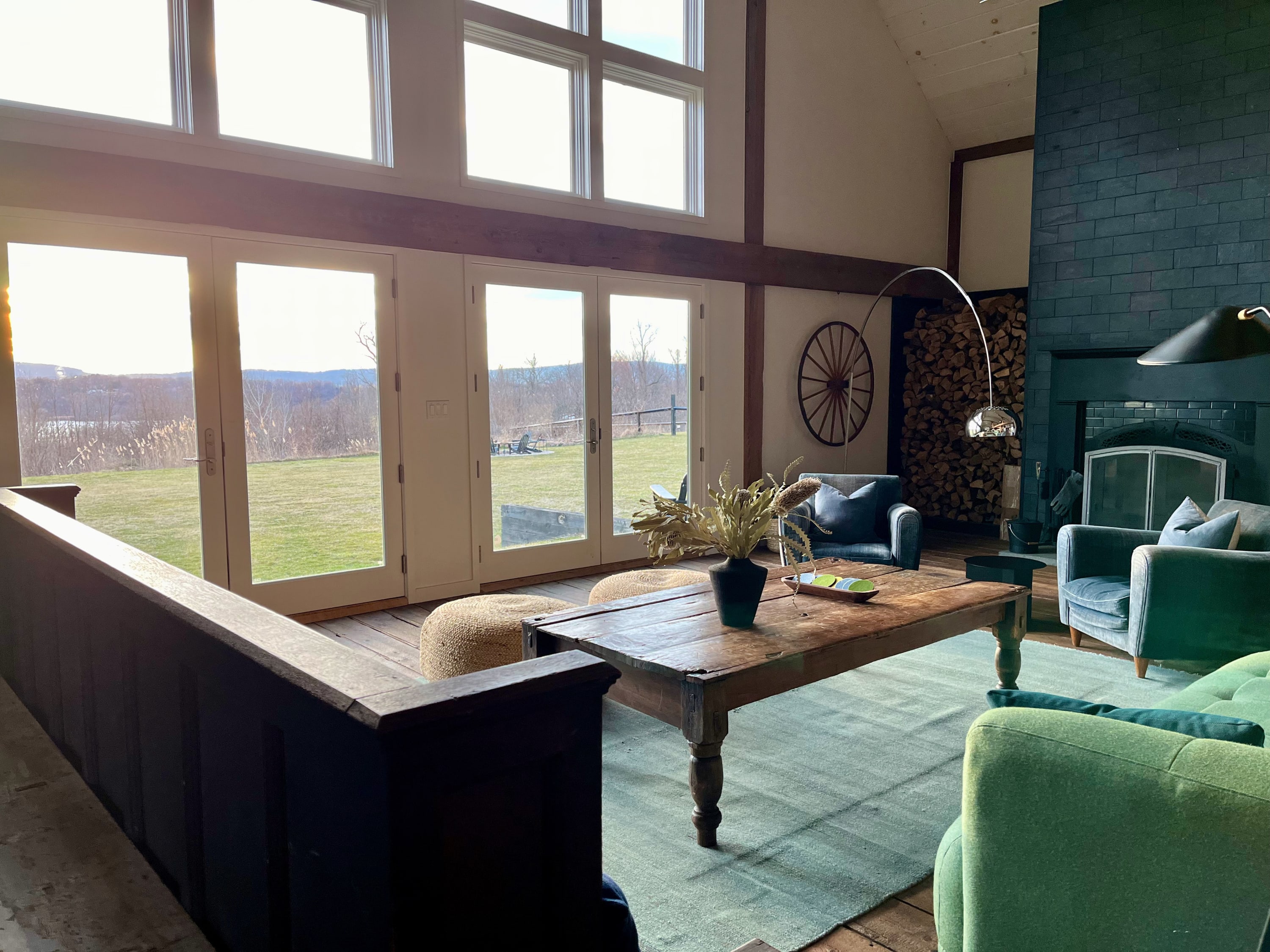 A bright living room boasts a vaulted ceiling with exposed beams, a large stone fireplace, and floor-to-ceiling windows offering expansive views of a green landscape. A rustic wooden coffee table sits on a light green rug, surrounded by various seating.