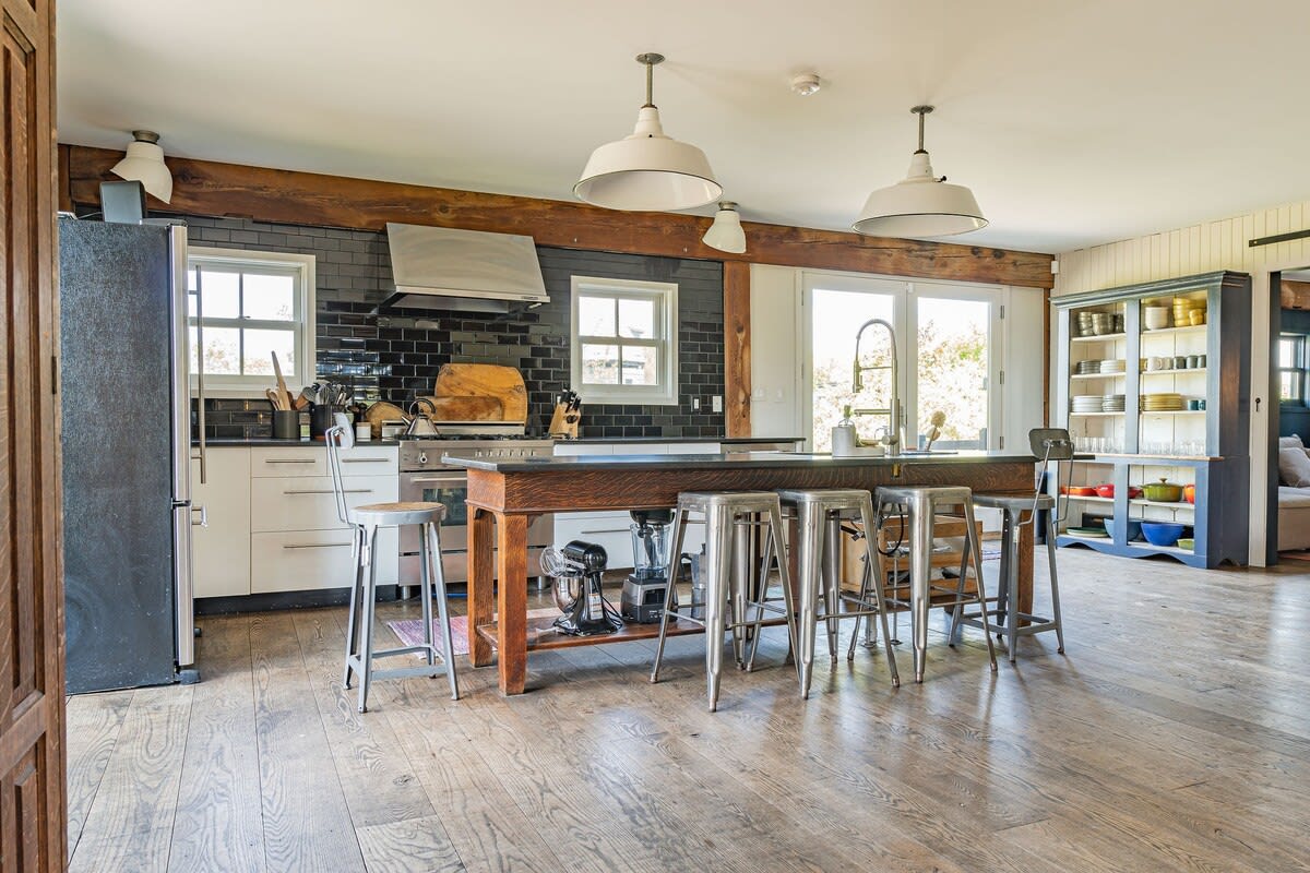 A bright, rustic-modern kitchen features a large wooden island with a dark countertop and metal bar stools, set against a black subway tile backsplash and white cabinetry. Exposed wooden beams cross the ceiling, complementing the wide-plank wood floors.
