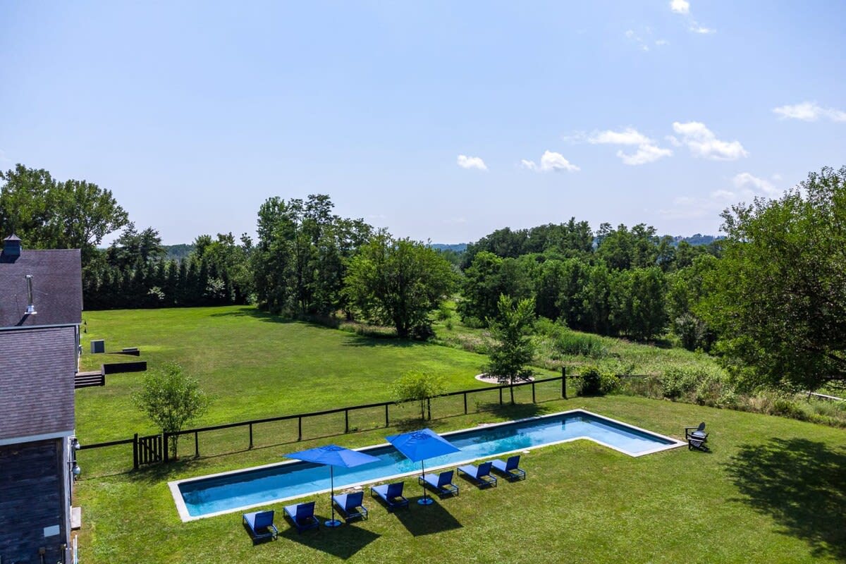 A wider aerial view showcasing the long, narrow swimming pool with blue lounge chairs and umbrellas, set within a large green lawn next to a dark barn-style house, with expansive green landscapes and distant mountains under a clear sky.