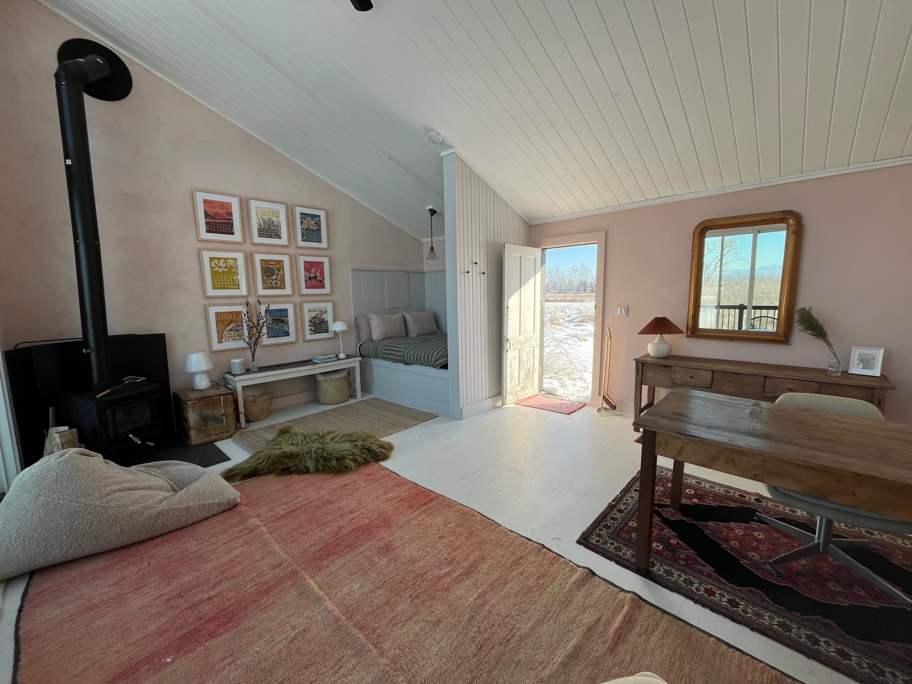 Spacious open-concept living area with light pink walls, white planked ceilings, a wood-burning stove, and a built-in bed nook, featuring multiple rugs and a wooden dining table, with an open door to a snowy exterior.