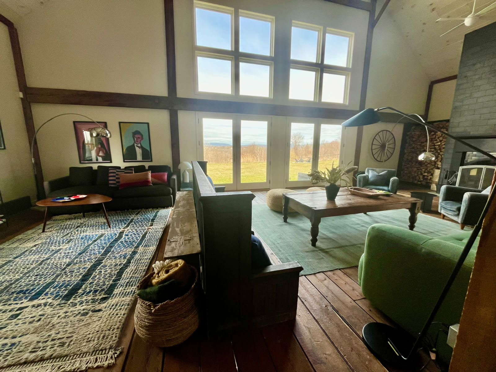 A bright and airy living room showcases a vaulted ceiling with exposed beams, large windows and glass doors overlooking a scenic landscape, and a prominent stone fireplace. Two distinct seating areas with colorful rugs define the open space.