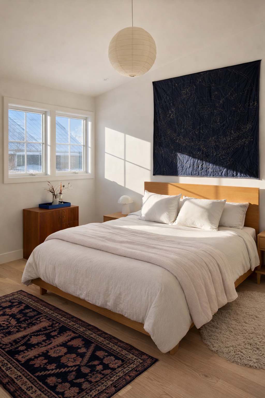 Bright and minimalist bedroom with a light wood bed frame, white bedding, and a large dark blue textile art piece above the headboard. Two windows let in ample natural light, illuminating the light wood floors and patterned rugs.