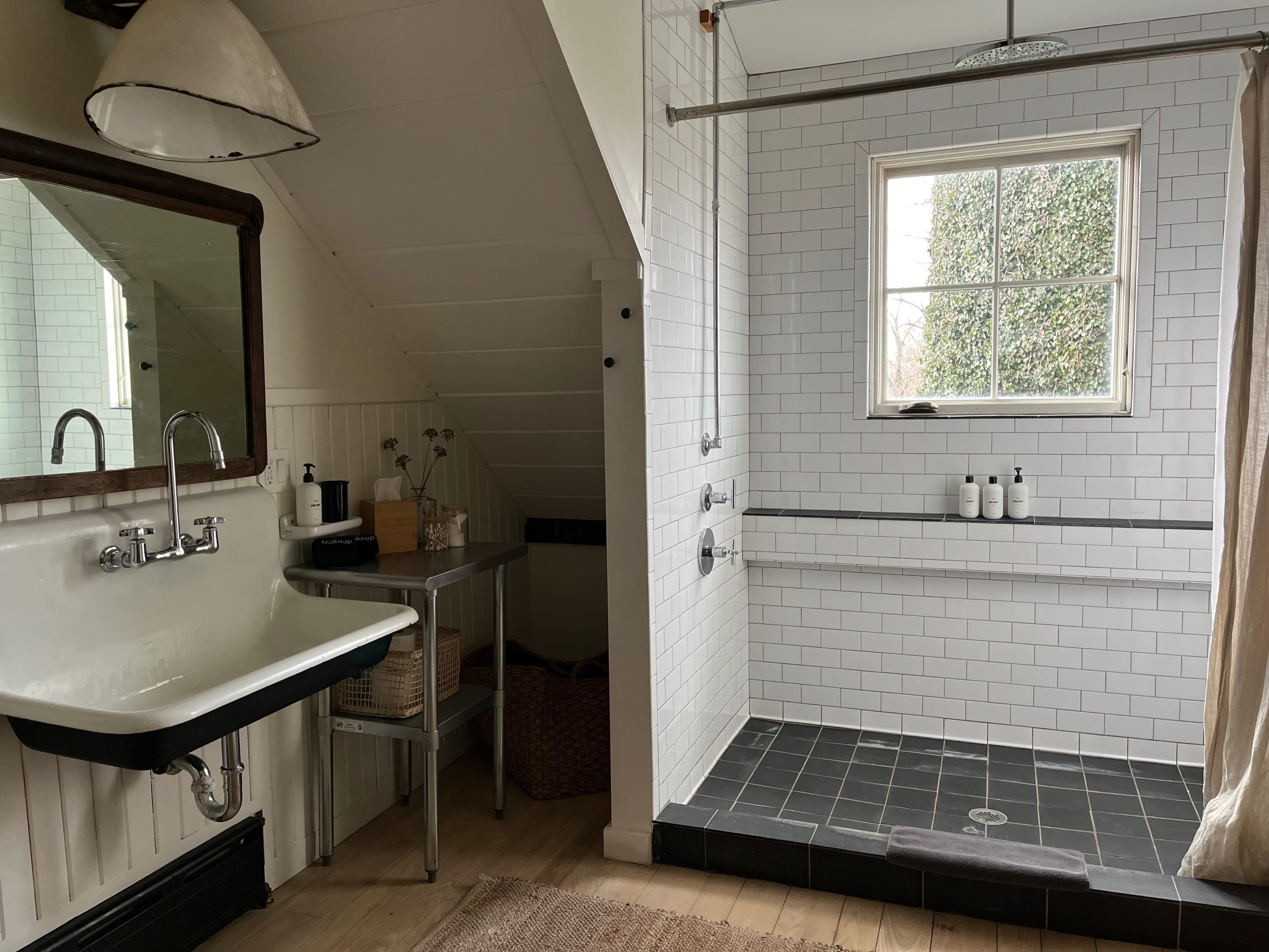 Another view of the bathroom, showing a vintage-style sink with a large mirror, a metal utility table, and the walk-in shower with subway tiles and a window.