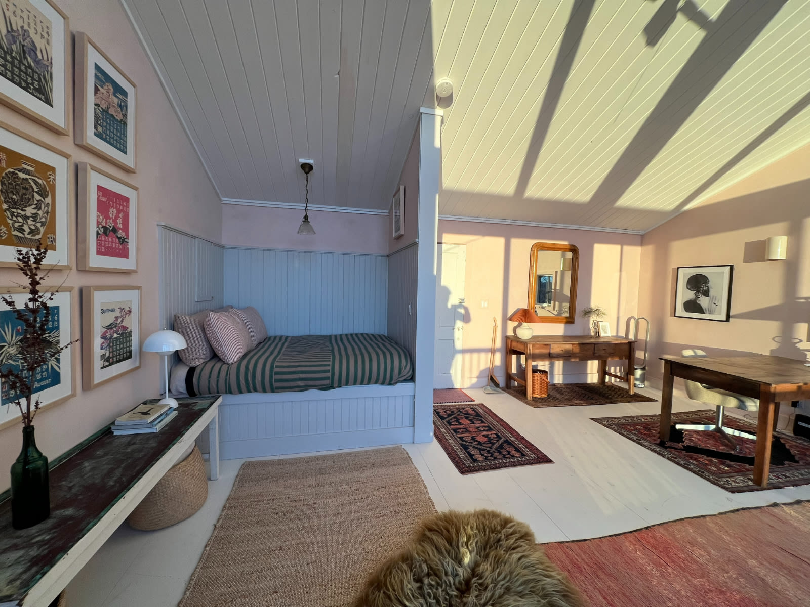 Open-concept living area with light pink walls and white planked ceilings, featuring a built-in bed nook with striped bedding, a long wooden console table, and a dining table, all bathed in warm sunlight.