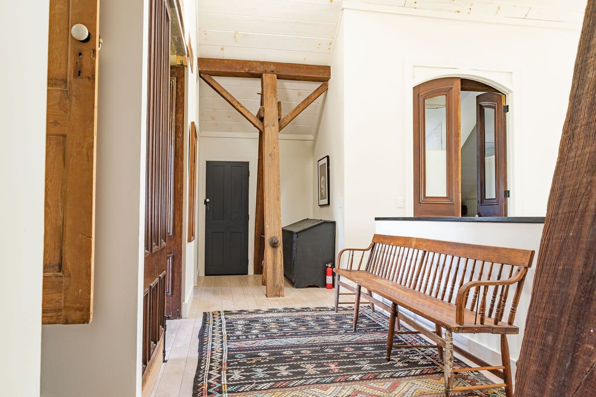 A rustic foyer features a long wooden bench, a patterned black and white rug, and prominent exposed wooden beams, with a dark door visible in the background.