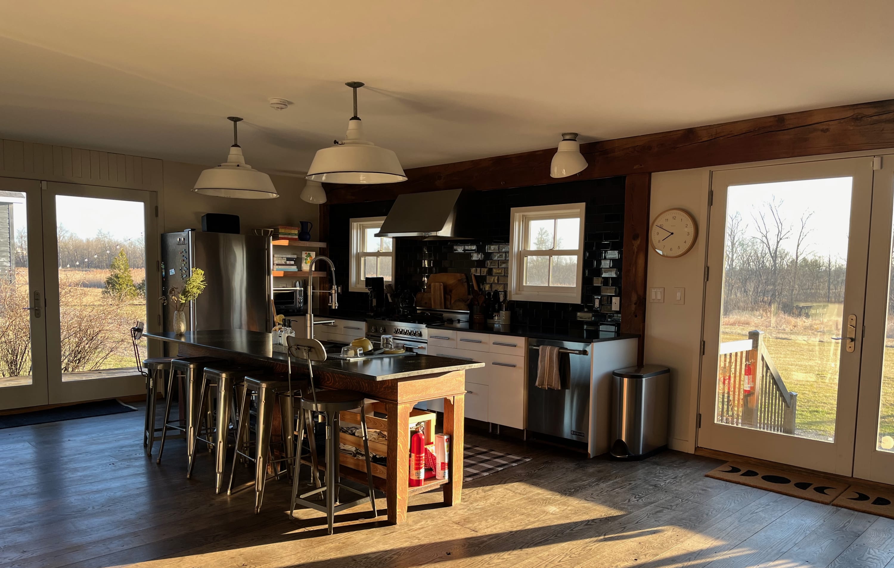 A sunlit, spacious kitchen with a large dark-topped wooden island and metal bar stools, surrounded by light wood floors. White cabinetry and a black subway tile backsplash line the wall, with large windows and glass doors offering views of the outdoors.