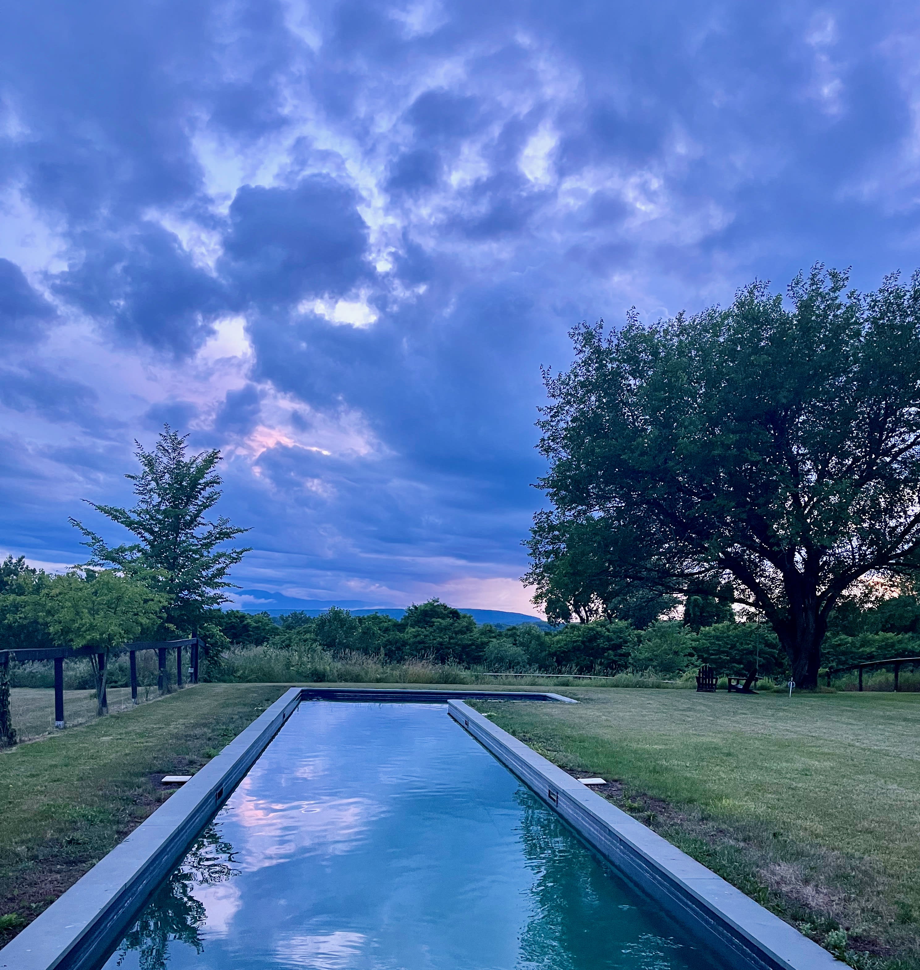 The long, narrow swimming pool reflects the dramatic purple and blue hues of a cloudy sunset sky, surrounded by green grass and trees, with distant mountains visible under the twilight.