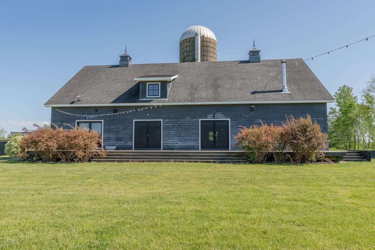The back of the dark gray barn-style house features multiple large doors and windows, with a raised wooden deck and decorative string lights, overlooking a green lawn and distant trees.