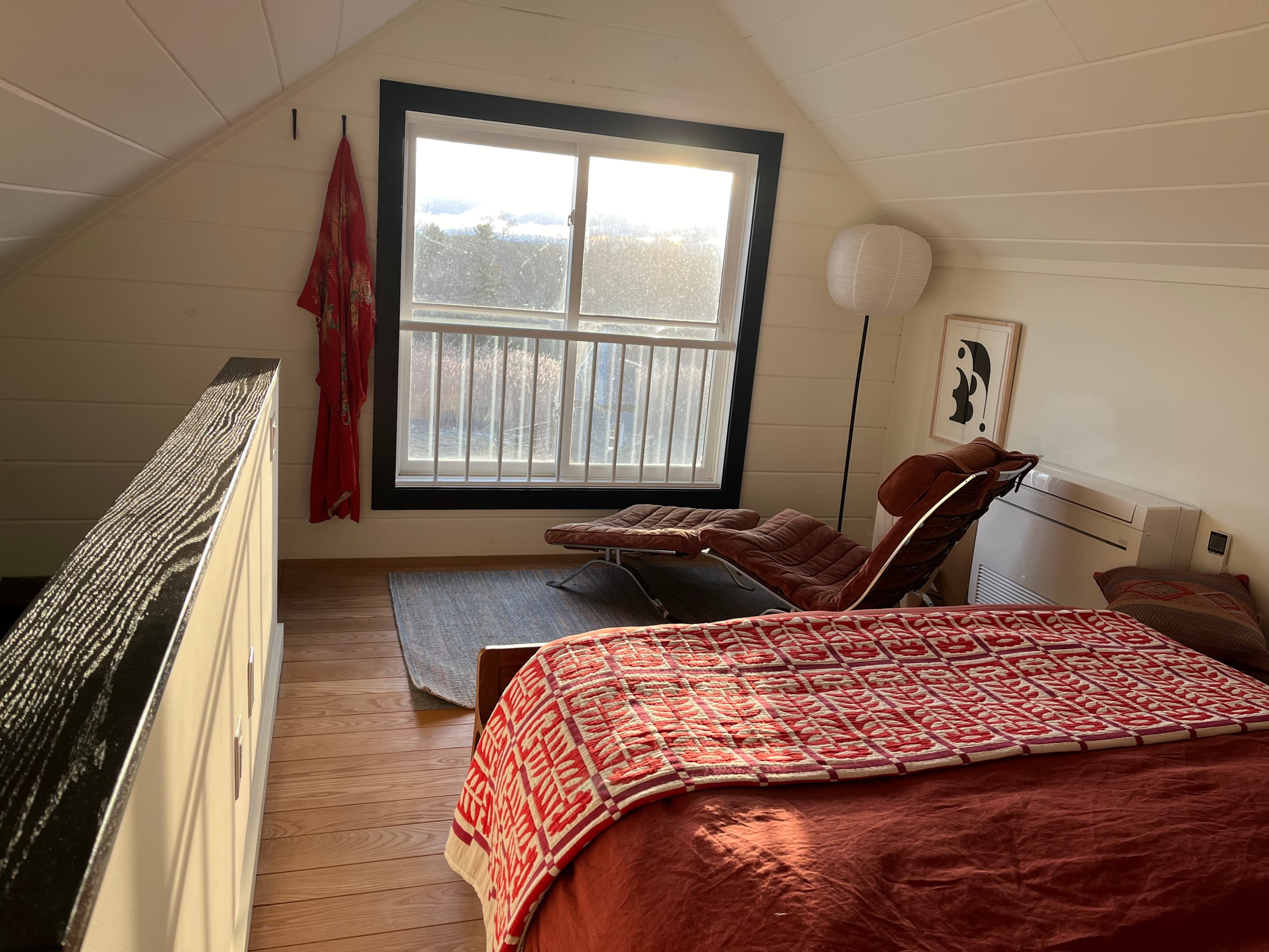 Another perspective of the bright bedroom, highlighting the large window with a black frame and a red kimono hanging nearby. The velvet lounge chair and ottoman are visible, along with a paper lantern floor lamp.