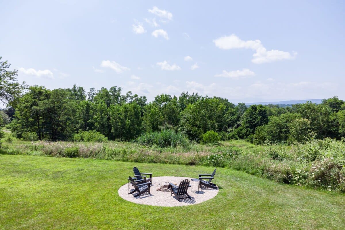 Outdoor fire pit area with four Adirondack chairs arranged around a gravel circle, set on a green lawn with a backdrop of dense green trees and distant rolling hills under a partly cloudy sky.