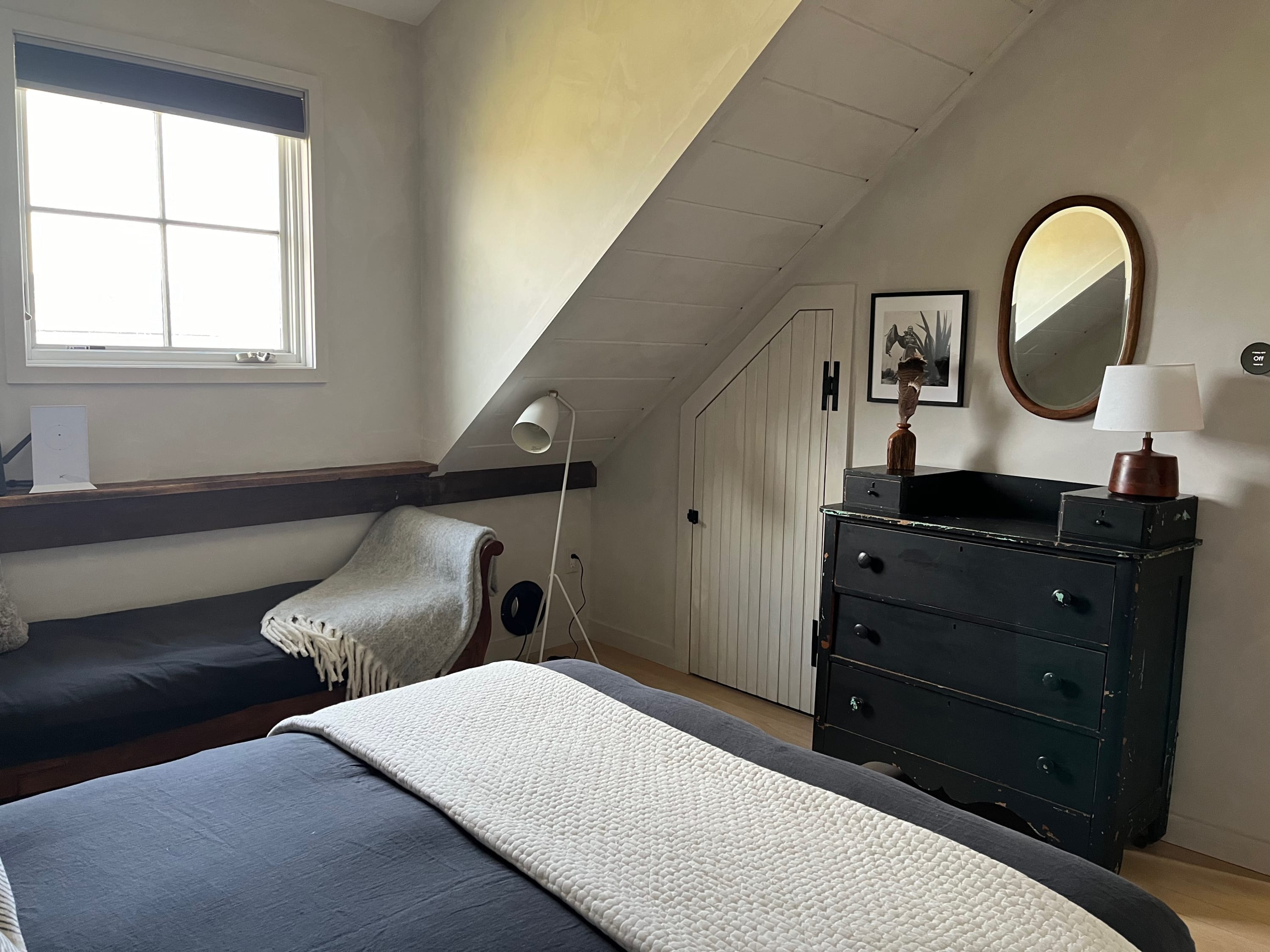 Another view of the cozy bedroom, featuring a dark wooden dresser with a round mirror above it. The slanted ceiling continues, and a small white door is visible under the eaves.