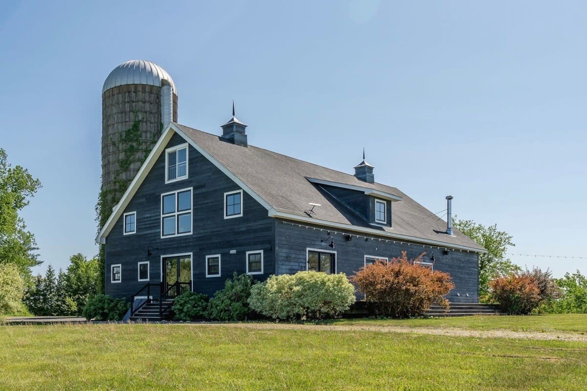 Ground-level view of the dark blue barn-style house with white trim and multiple windows, adjacent to a tall concrete silo covered in ivy, set on a green lawn under a clear blue sky.
