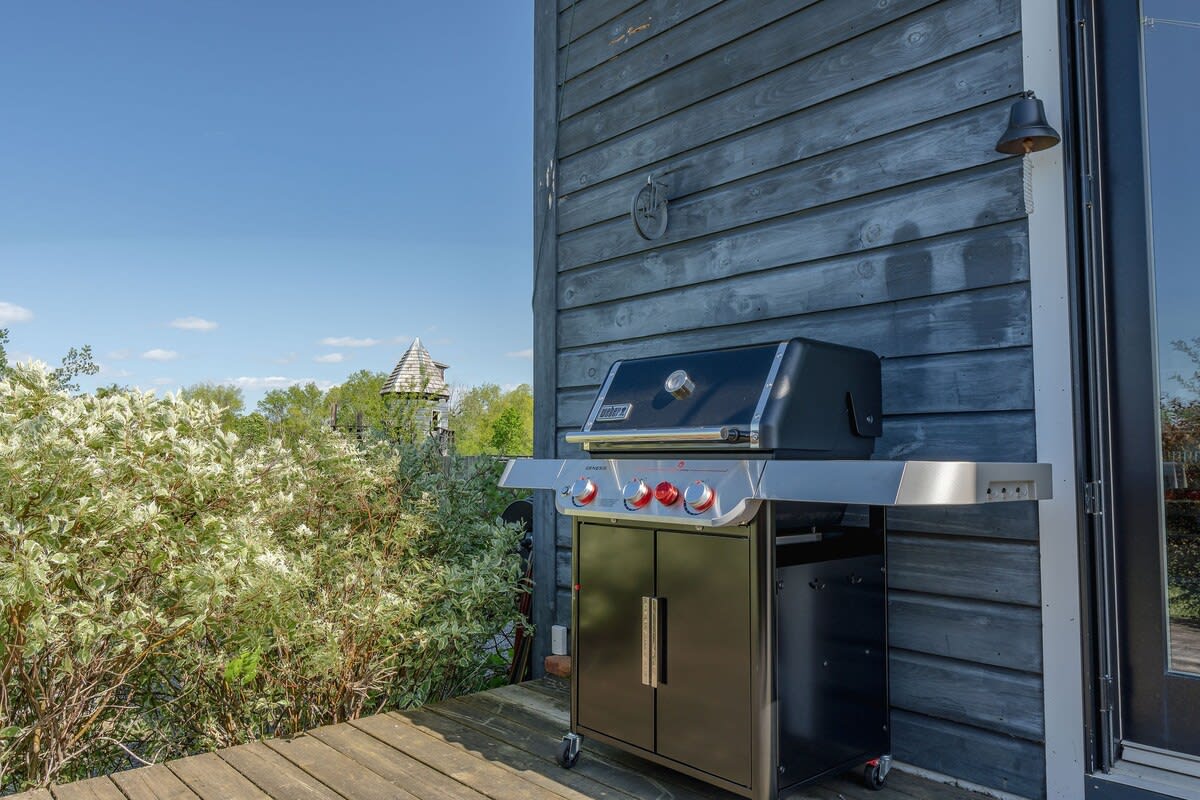 A black gas grill with red knobs sits on a wooden deck next to a dark gray wooden barn-style building, surrounded by lush green and white flowering bushes under a clear blue sky. A small bell hangs on the wall above the grill.