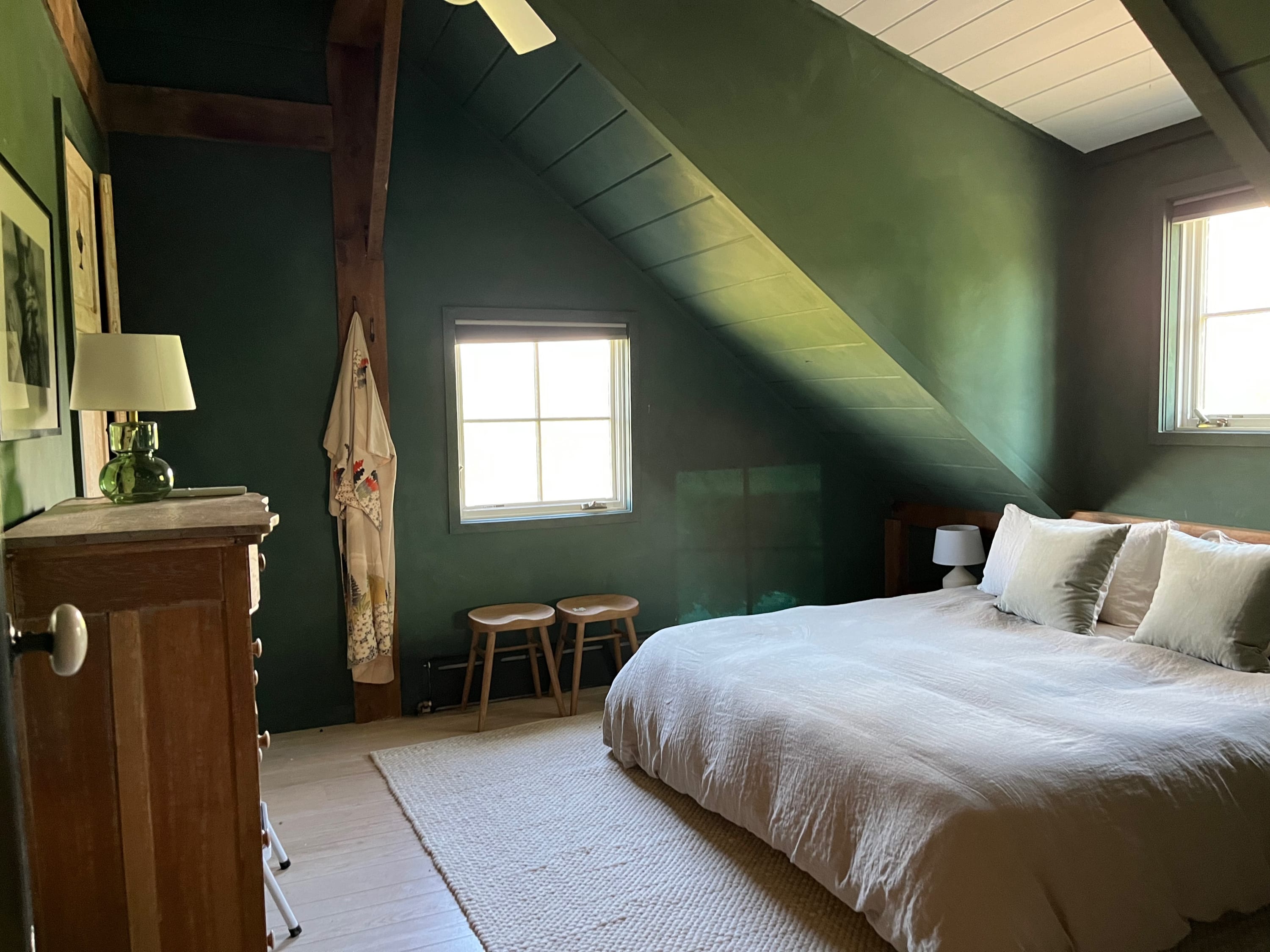 Another view of the green-walled bedroom, highlighting the bed with light bedding, a wooden dresser with a lamp, and two small wooden stools beneath a window, emphasizing the sloped ceiling.