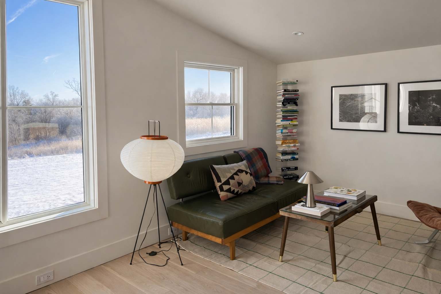 Cozy living area with light hardwood floors, a green leather sofa, a glass-top coffee table, and a tall paper lantern floor lamp, offering views of a snowy landscape through large windows.
