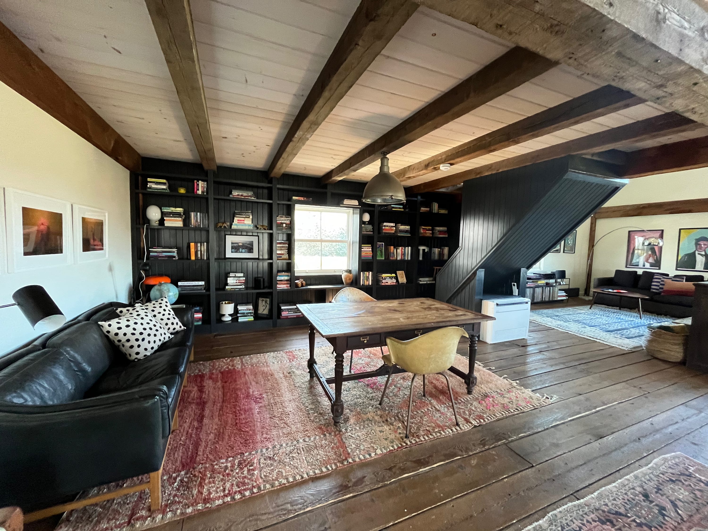 A cozy living space features a wall-to-wall black built-in bookshelf filled with books, a rustic wooden table with a yellow chair, and a black leather sofa, all on a patterned red rug. Exposed ceiling beams add to the rustic charm.