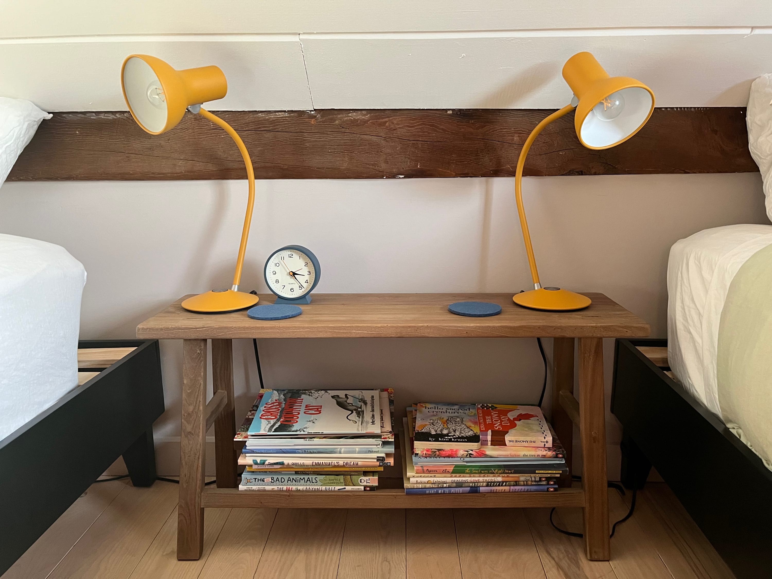 A close-up of the wooden nightstand between the two twin beds, featuring two yellow desk lamps, a blue alarm clock, and stacks of books on the lower shelf.