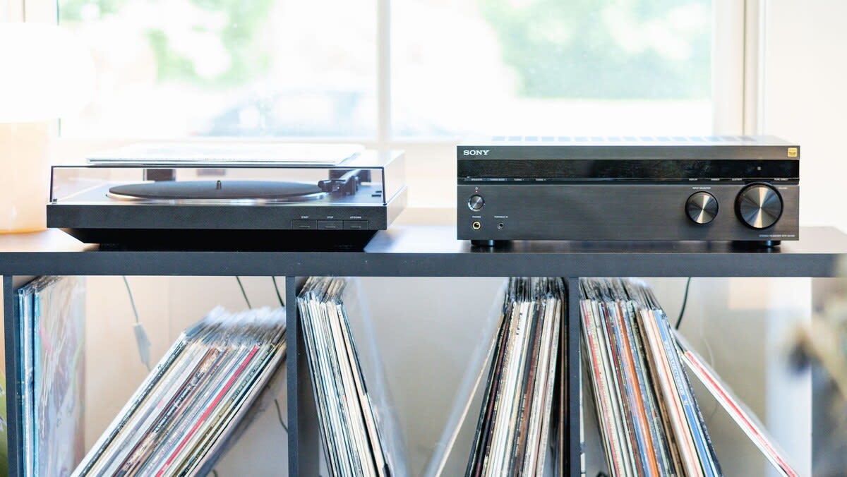 A close-up view of a modern record player and a Sony audio receiver, neatly arranged on a black shelf above a collection of vinyl records. The setup is positioned near a window, allowing natural light.