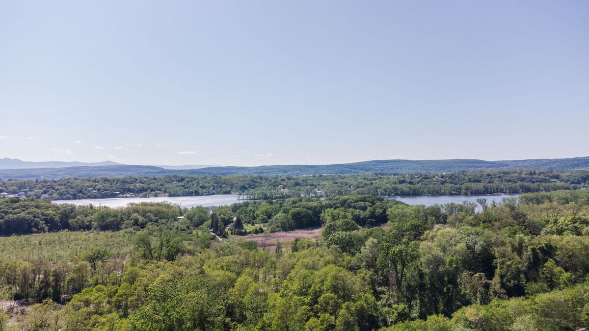 A panoramic aerial view of a vast green landscape with dense forests, a winding river or lake, and distant mountains under a clear blue sky, highlighting the property's scenic surroundings.