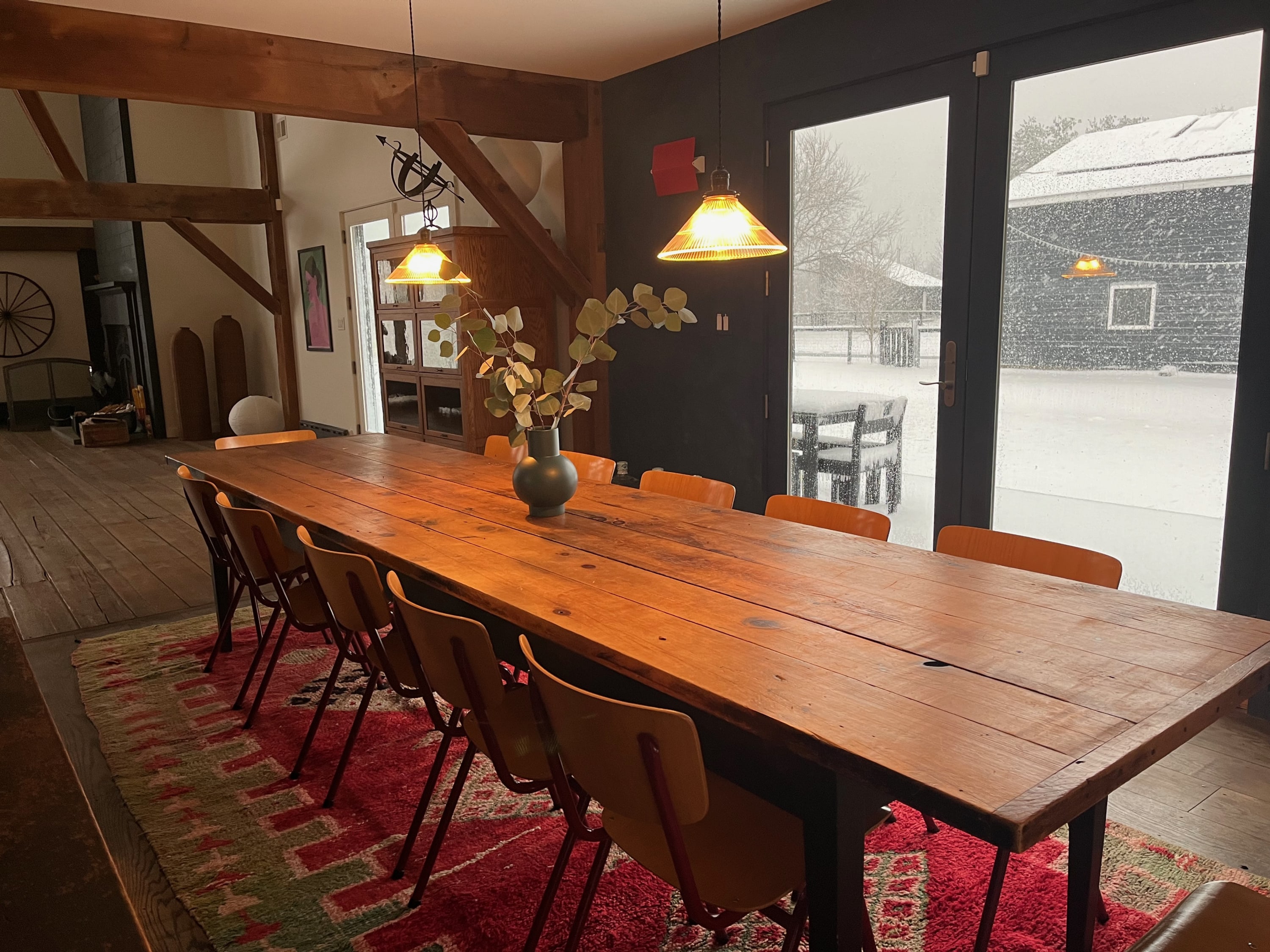 A spacious dining room features a long, rustic wooden dining table surrounded by numerous orange chairs, set on a vibrant red and green patterned rug. Large glass doors offer a view of a snowy outdoor scene.