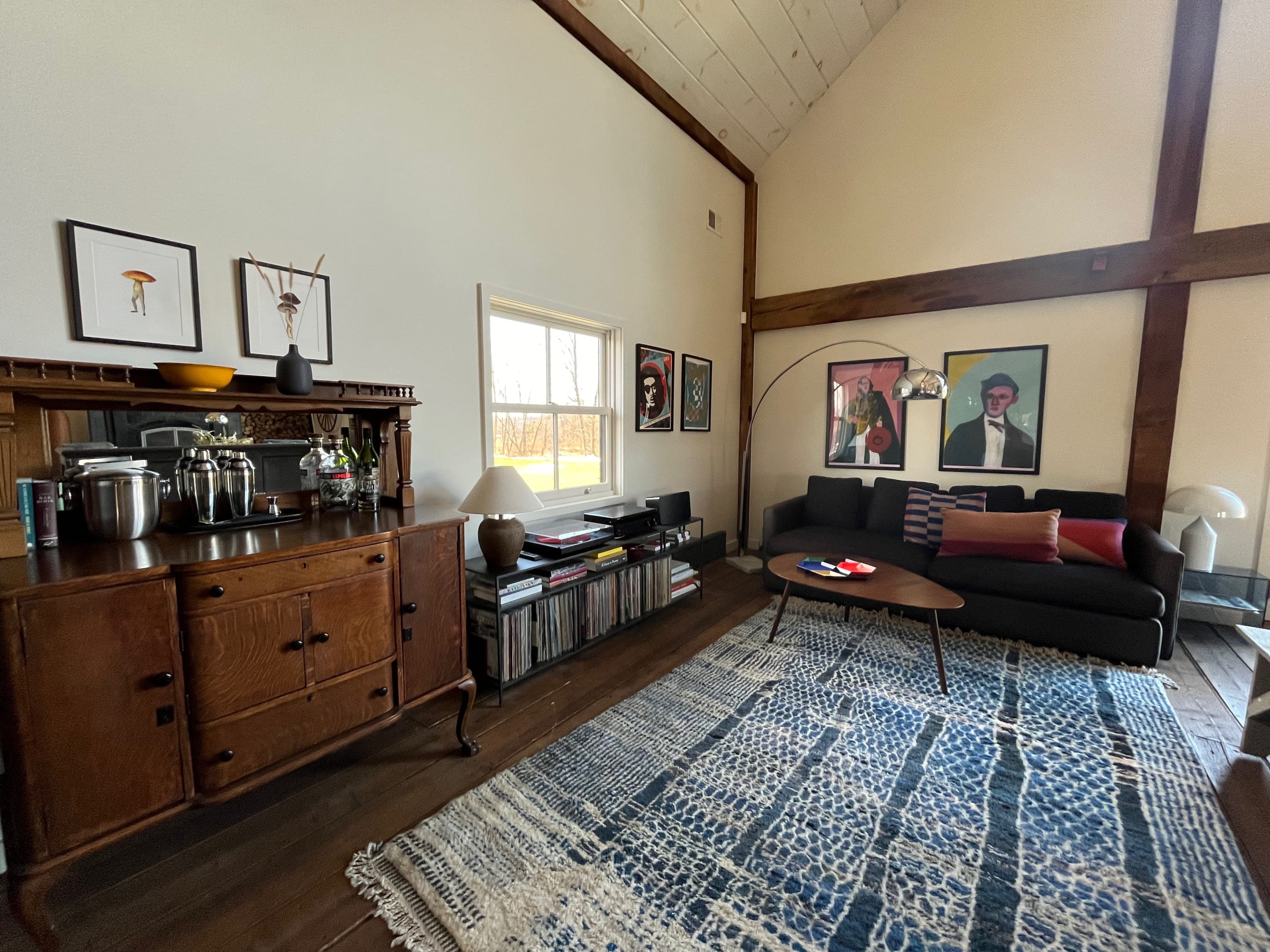 A stylish living room features a vintage wooden bar cabinet with barware, a record player setup on a shelf filled with vinyl records, and a dark sofa with colorful pillows, all on a blue and white patterned rug. Artwork adorns the white walls.
