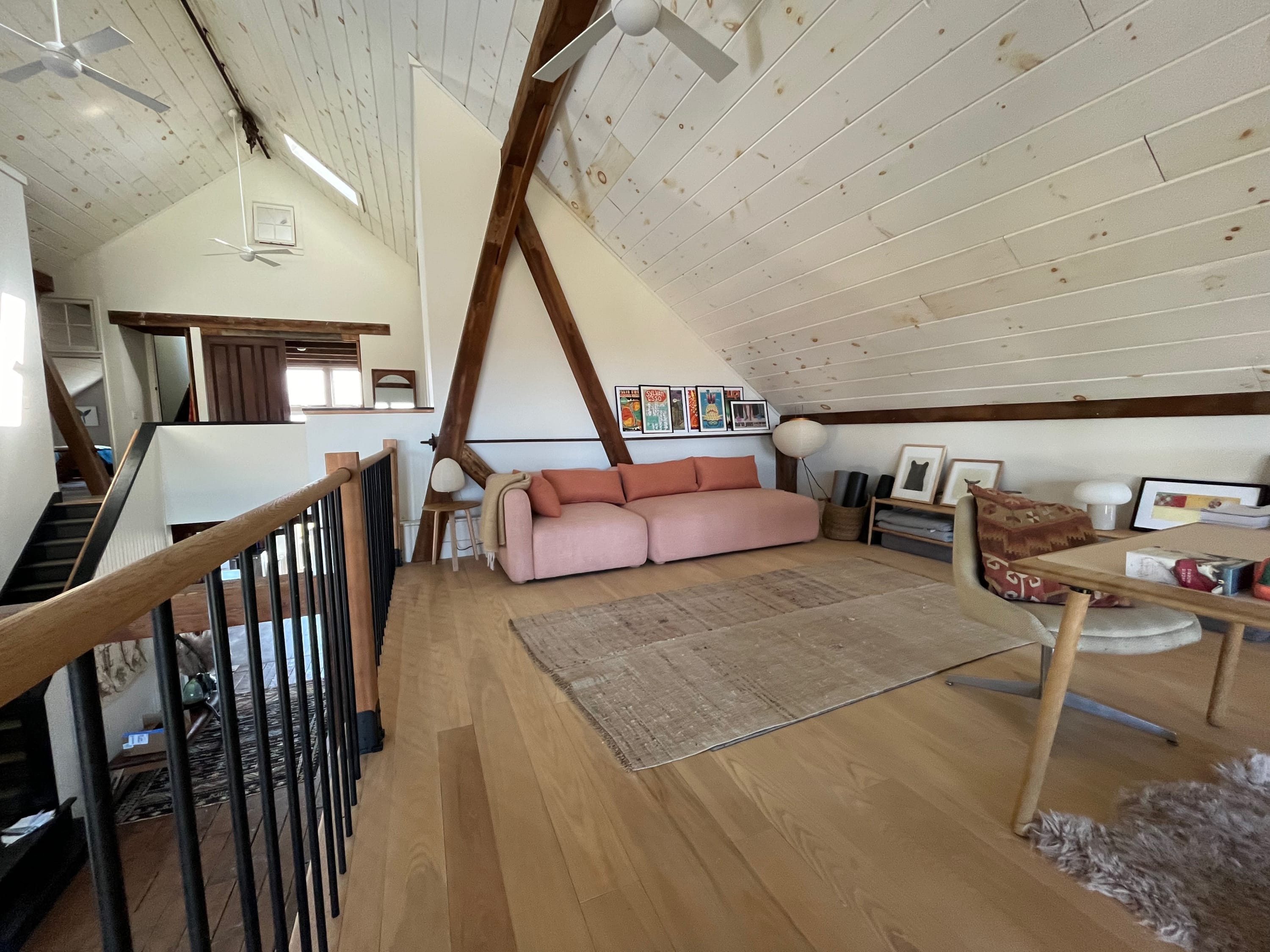 Expansive loft living area featuring a long pink sofa, a large textured rug, and exposed wooden beams. The vaulted, slatted white ceiling and open railing create an airy, bright space with views of the lower level.