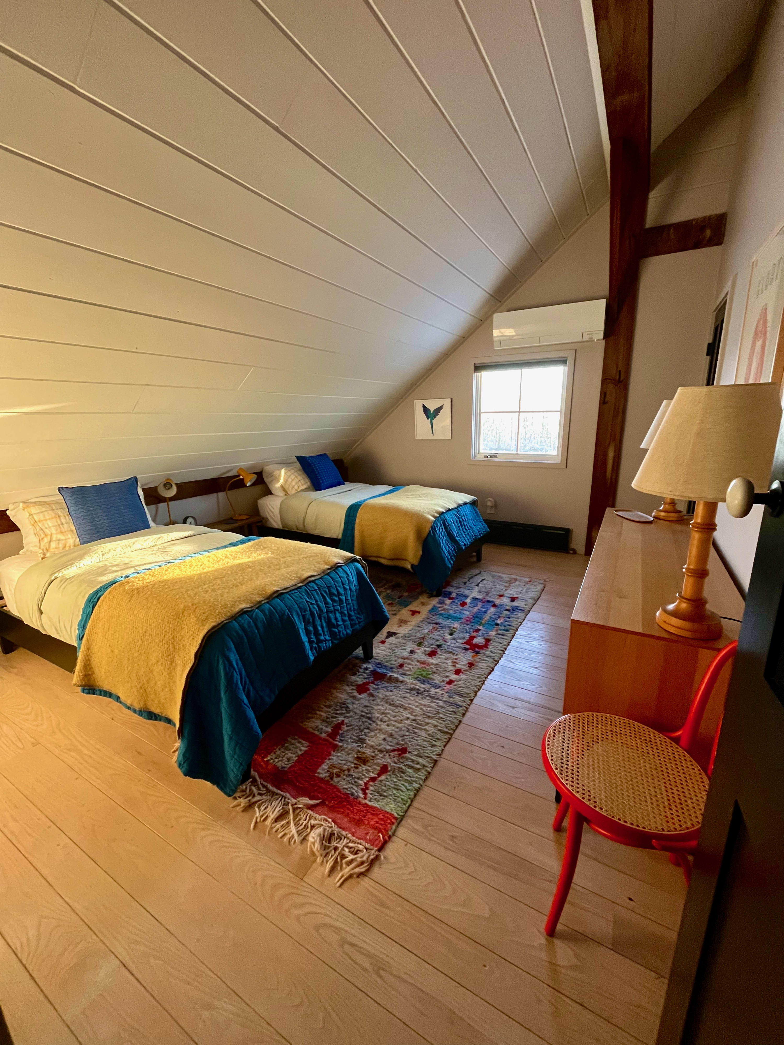 Another perspective of the twin bedroom, showing both beds with their vibrant bedding, a wooden dresser with a lamp, and a red chair, all under a sloped white ceiling.