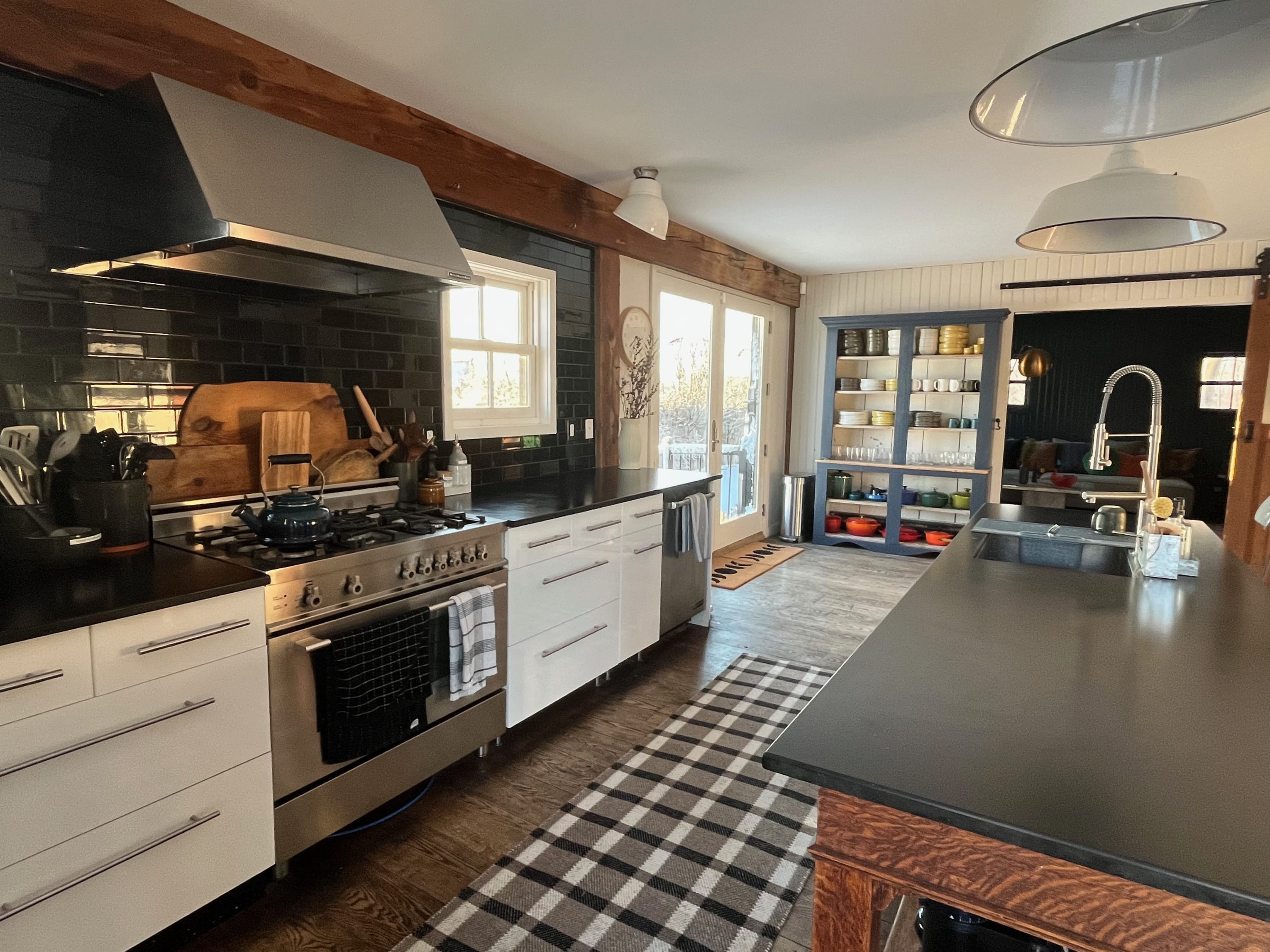 A close-up view of the rustic-modern kitchen highlights the black subway tile backsplash, stainless steel range, and white lower cabinets. A large wooden island with a dark countertop and sink is visible, along with a black and white checkered rug on the wood floor.