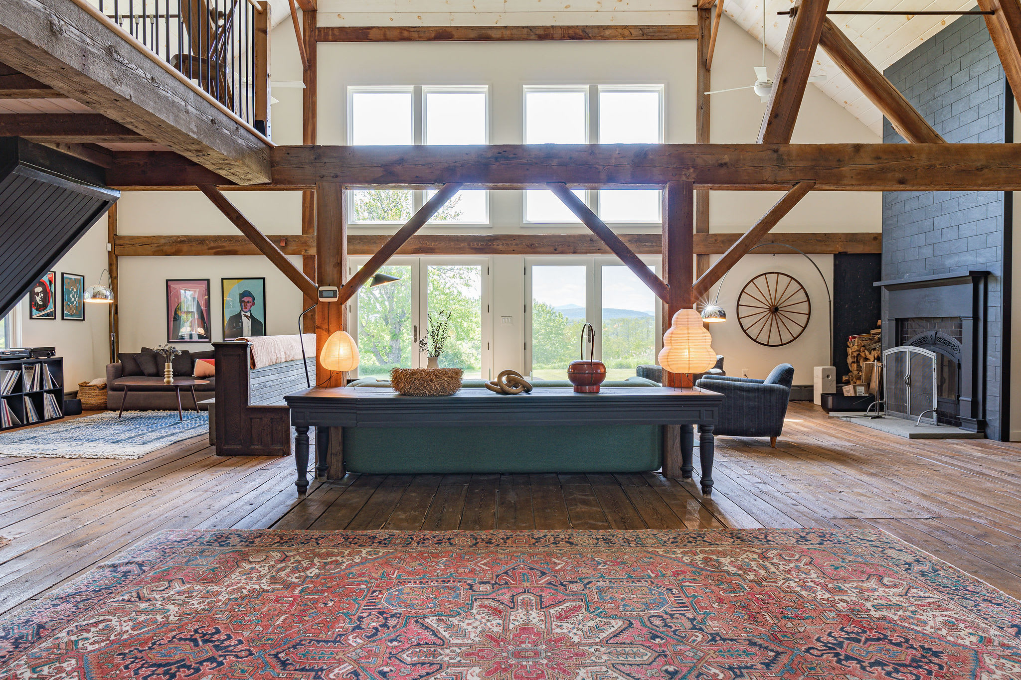 Expansive open-plan living area with high ceilings, exposed wooden beams, and wide plank wooden floors, featuring a large dark fireplace, a long wooden console table, and a seating area with a sofa and artwork.