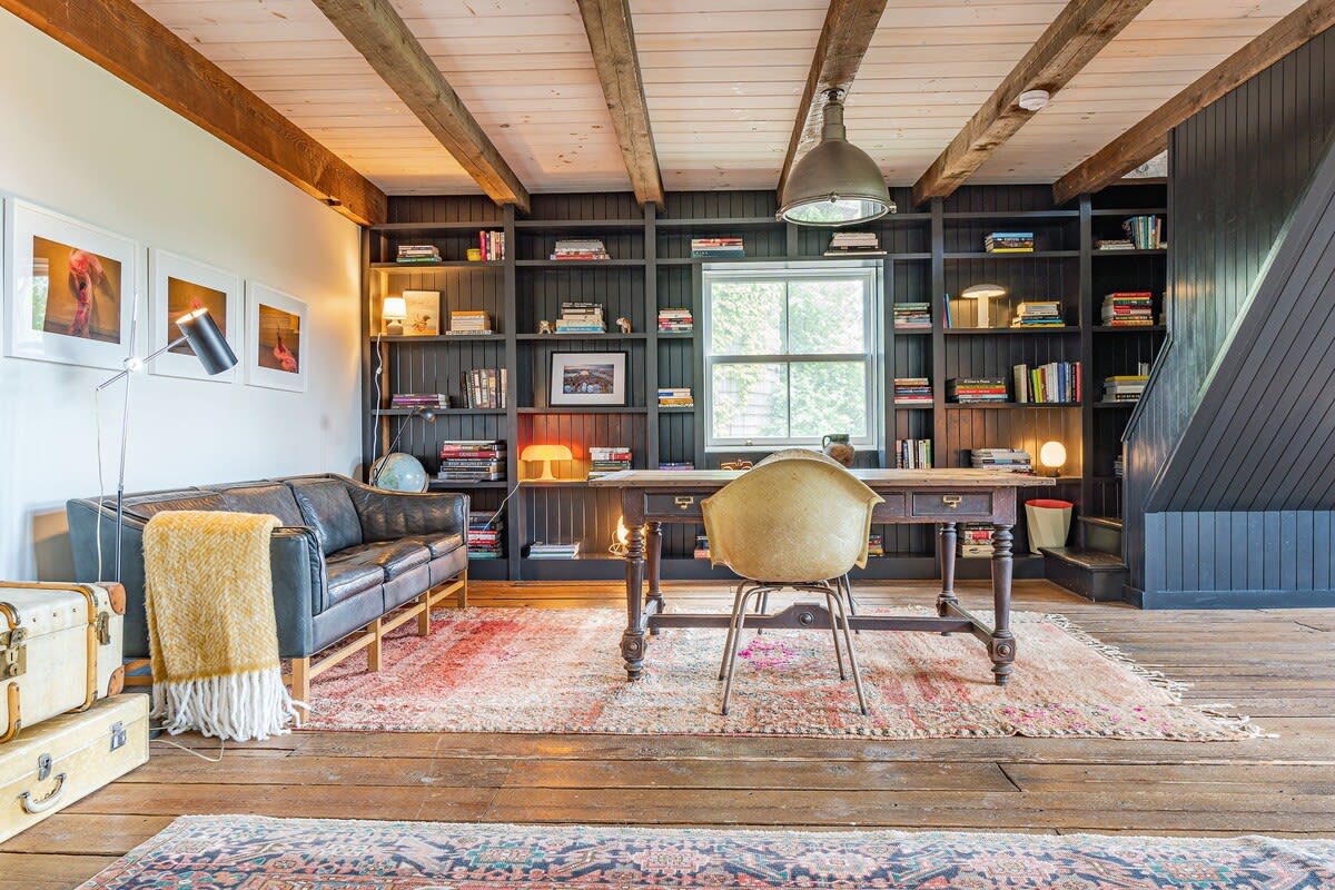 Rustic home office featuring dark built-in bookshelves filled with books, a large wooden desk with a vintage chair, a black leather sofa, and exposed wooden ceiling beams, all on wide plank wooden floors.