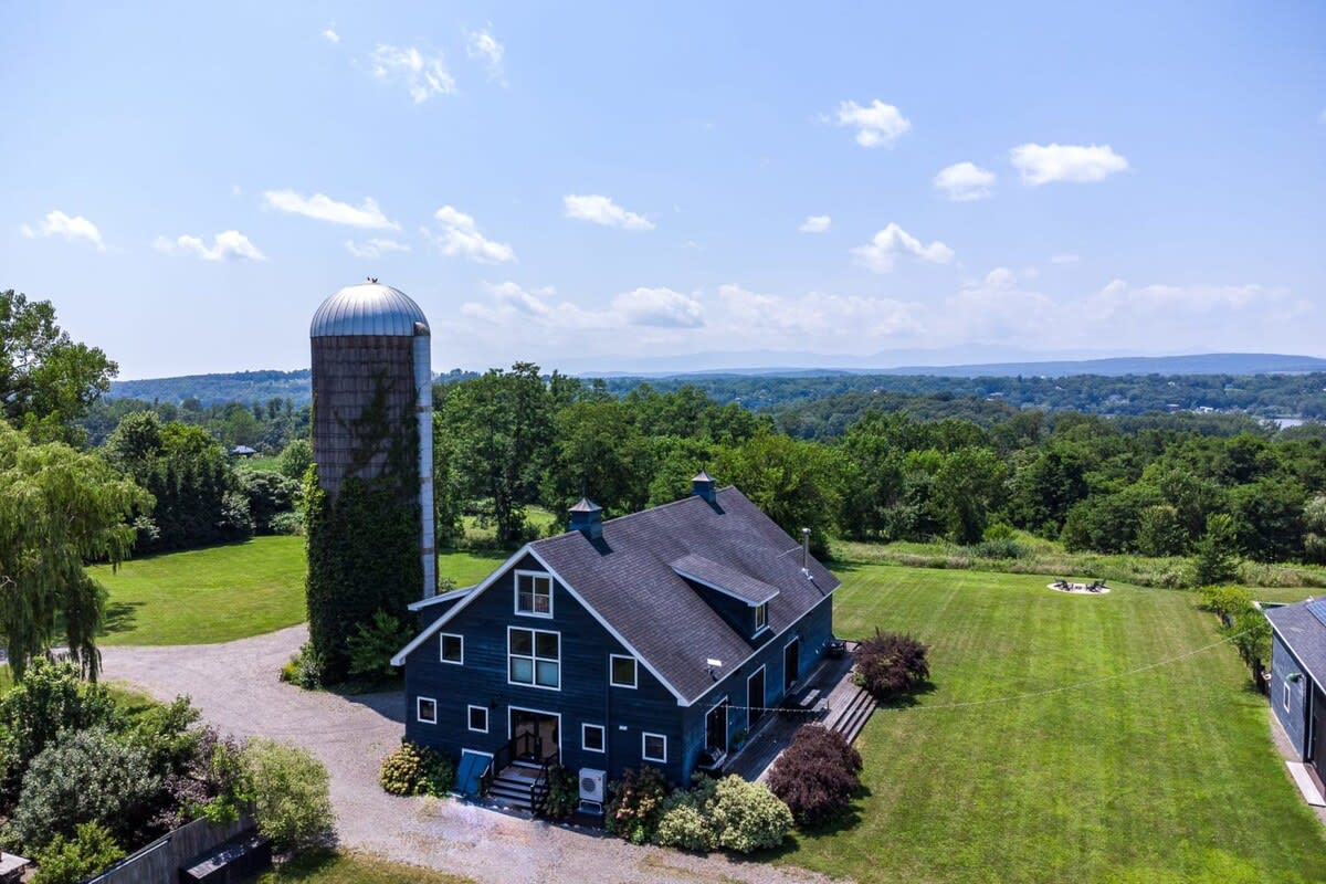 An aerial view of the dark gray barn-style house with a tall silo, surrounded by expansive green lawns and lush trees, with distant rolling hills and a body of water under a bright blue sky.