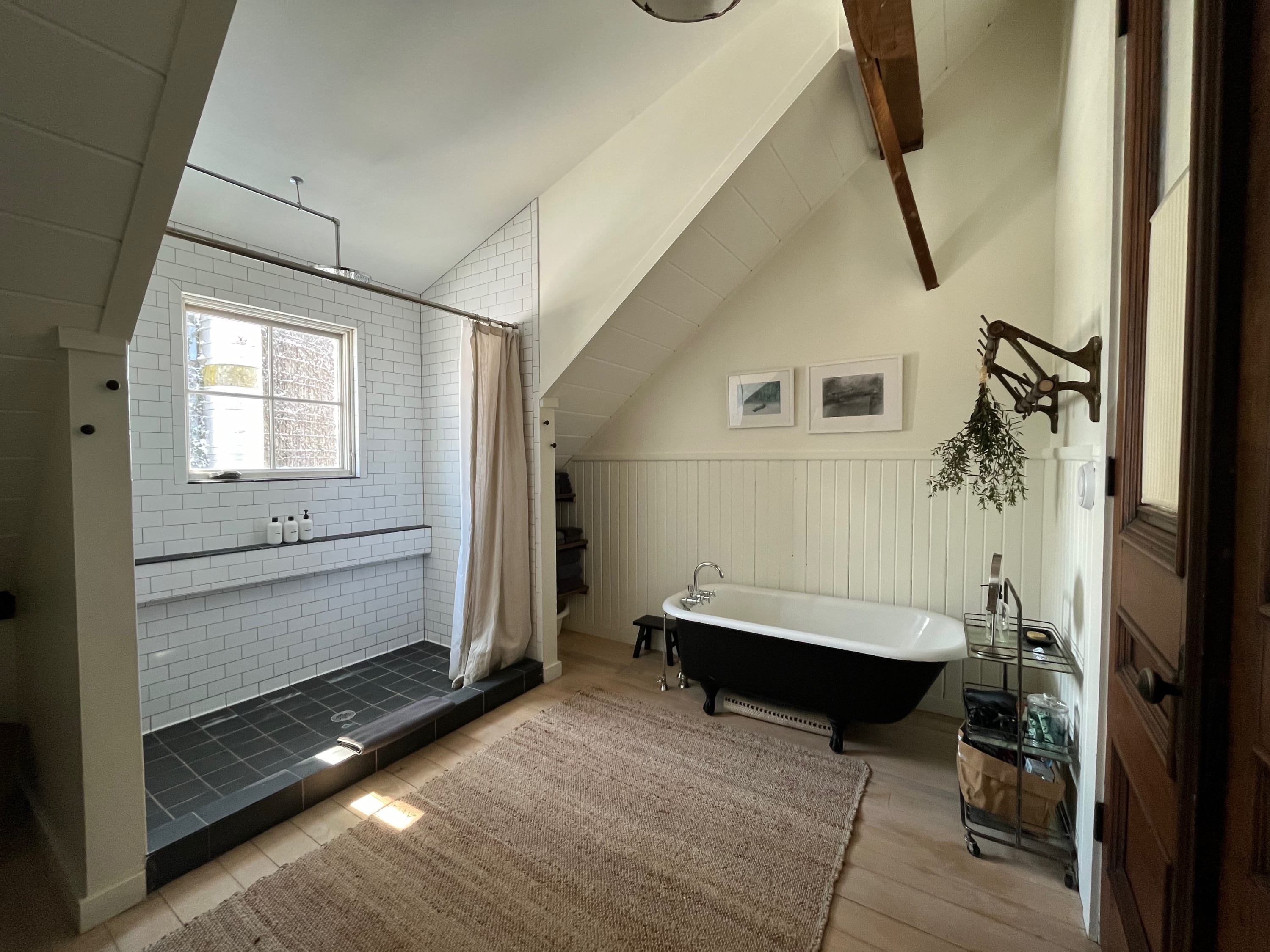 A bright bathroom with white paneled walls and a sloped ceiling features a spacious walk-in shower with subway tiles and a black base, alongside a classic black clawfoot tub.