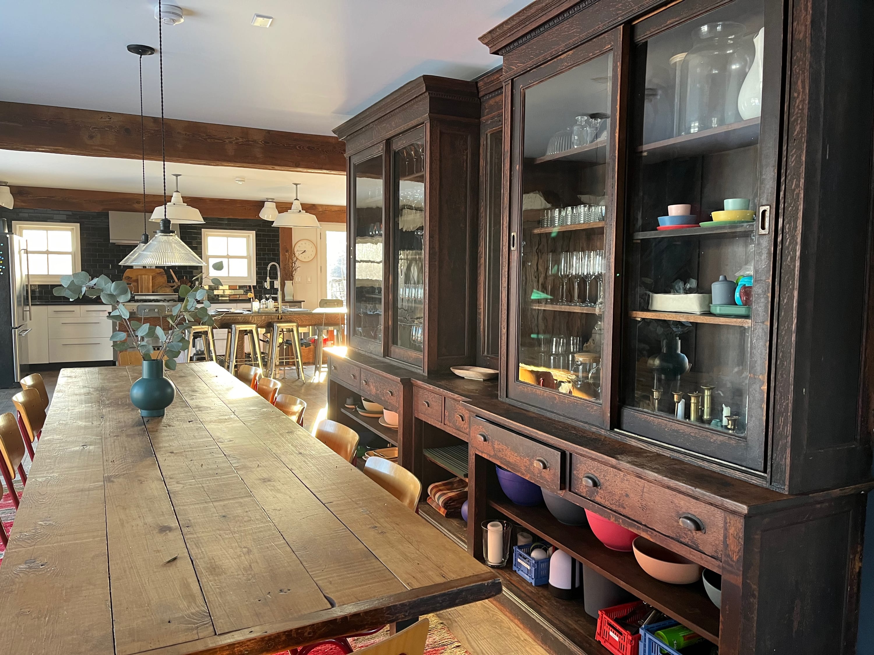 A long, rustic wooden dining table with a vase of greenery and colorful chairs anchors a spacious dining area, which seamlessly transitions into a modern kitchen visible in the background. Two large wooden display cabinets line the wall.