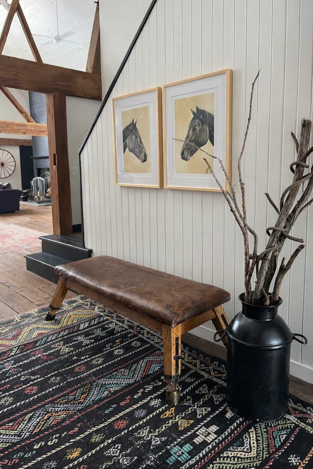 A rustic entryway features a vintage leather gymnastics bench, two framed horse head artworks on a white shiplap wall, and a large black metal vase filled with dried branches, all set on a colorful patterned rug.