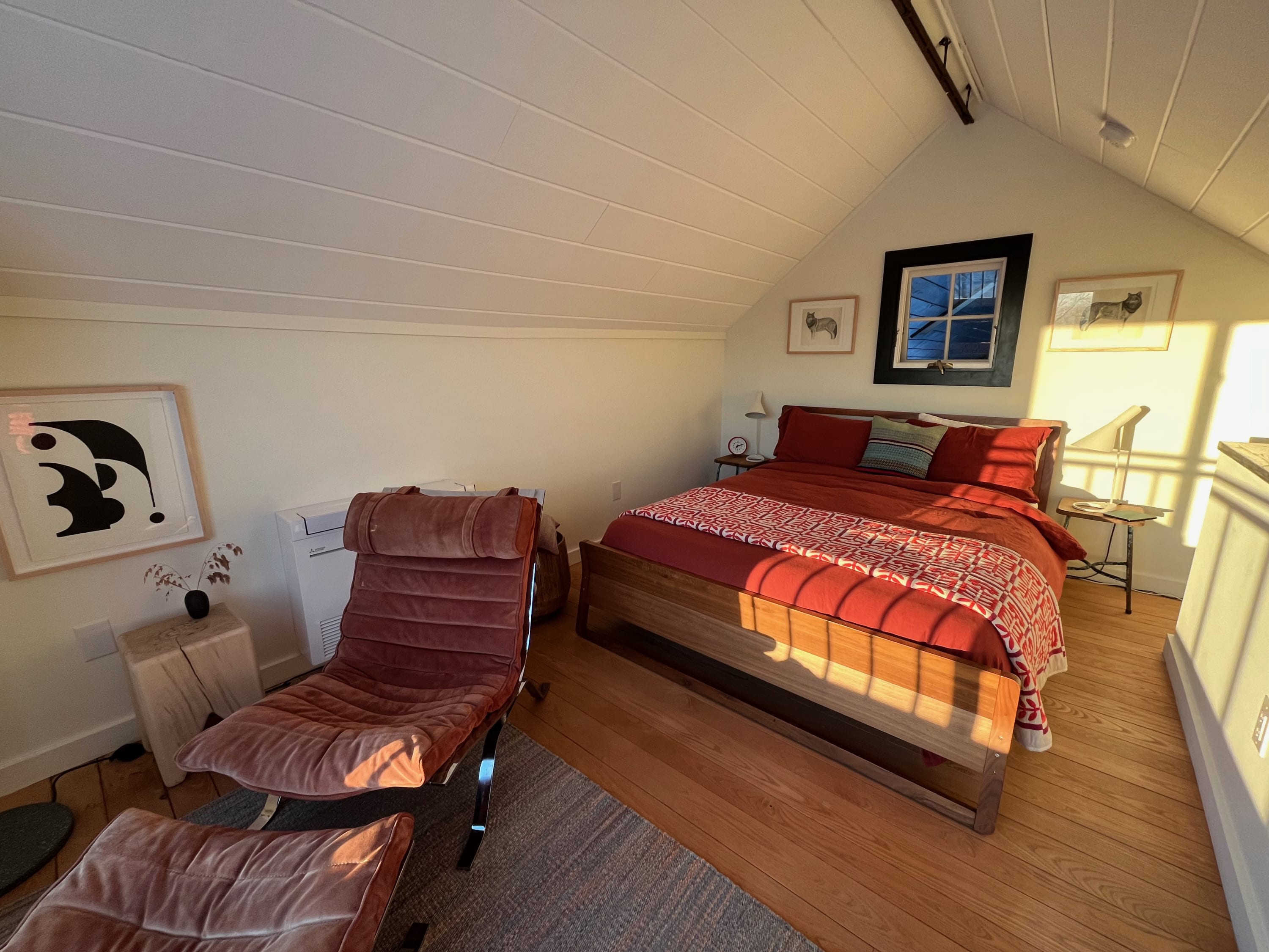 Bright bedroom with a wooden bed frame, red bedding, and a plush velvet lounge chair with an ottoman. Slanted white paneled ceilings and a large window with a black frame create a cozy, sunlit space.