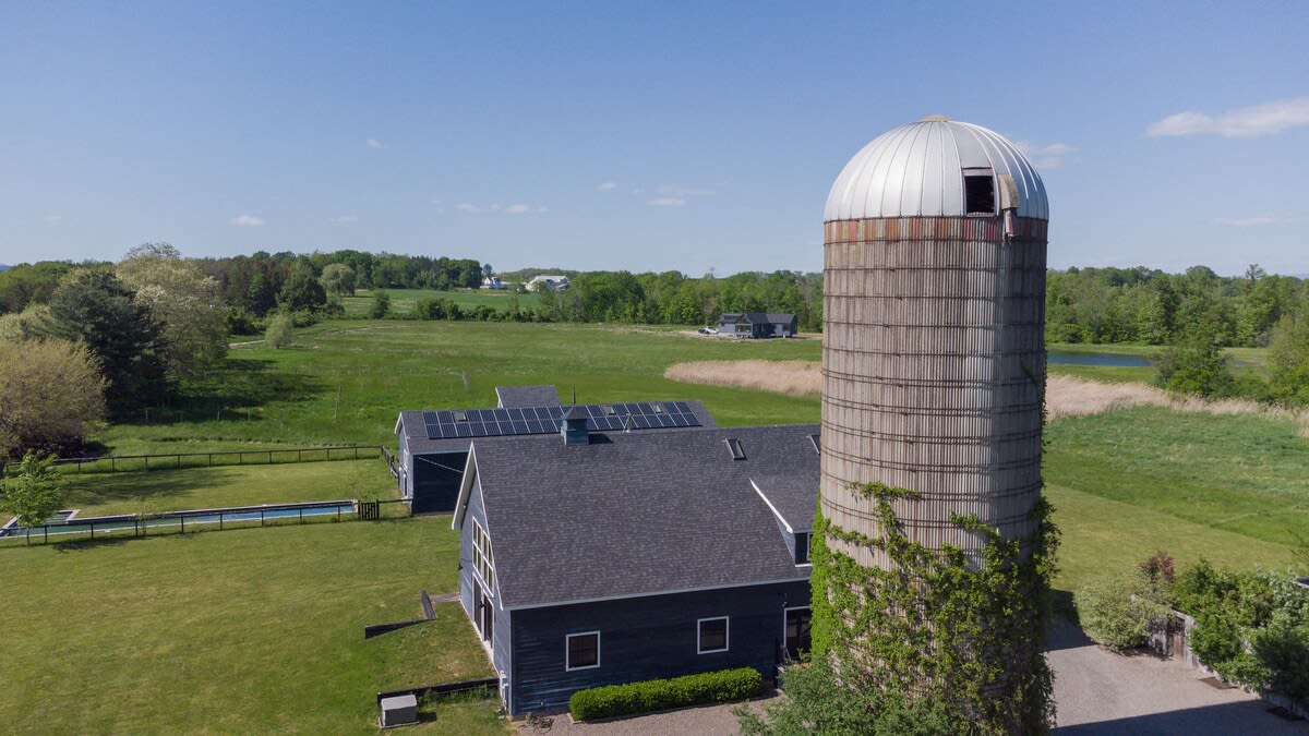 An aerial view showcases a dark blue barn-style house with solar panels on its roof, a prominent tall silo, and a long rectangular swimming pool, all nestled within expansive green fields and surrounded by lush trees under a bright blue sky.
