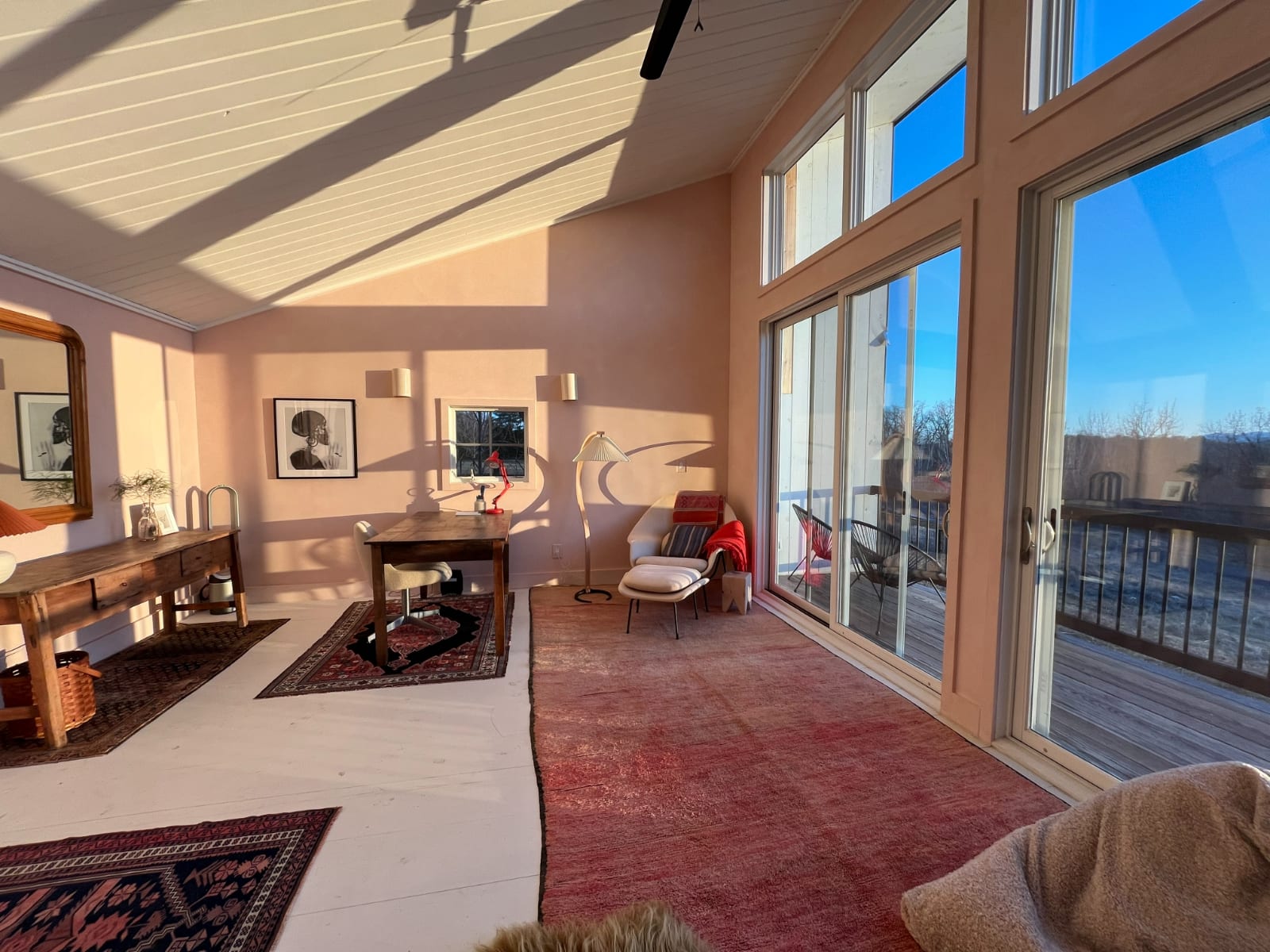 Bright living space with light pink walls and a high white planked ceiling, featuring large windows and sliding glass doors that open to a deck, showcasing a wooden dining table and various rugs.