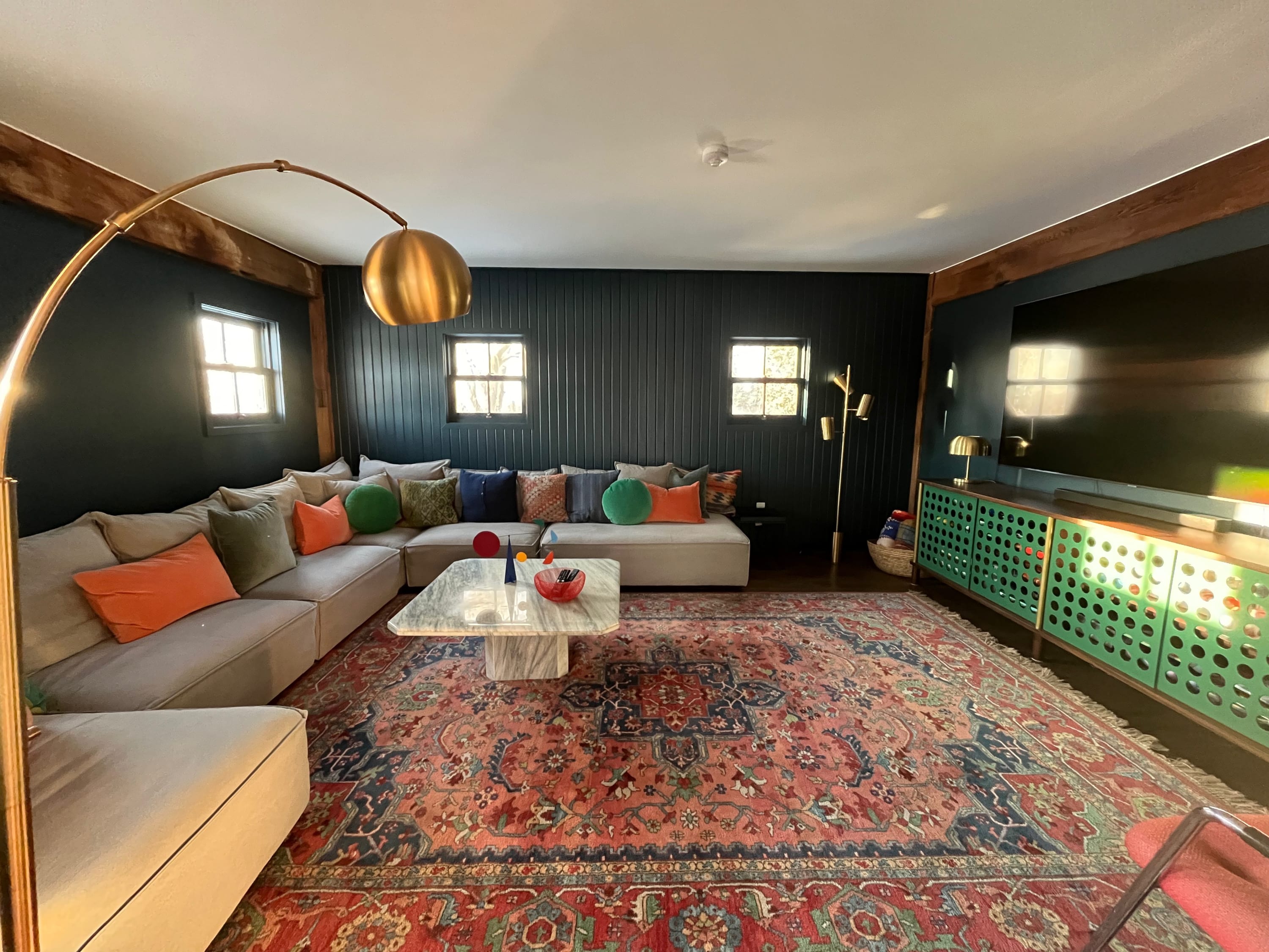 A cozy living room with dark paneled walls, a large L-shaped sectional sofa adorned with colorful pillows, and a marble coffee table. A vibrant patterned rug covers the wood floor, and a green cabinet with a large TV completes the space.