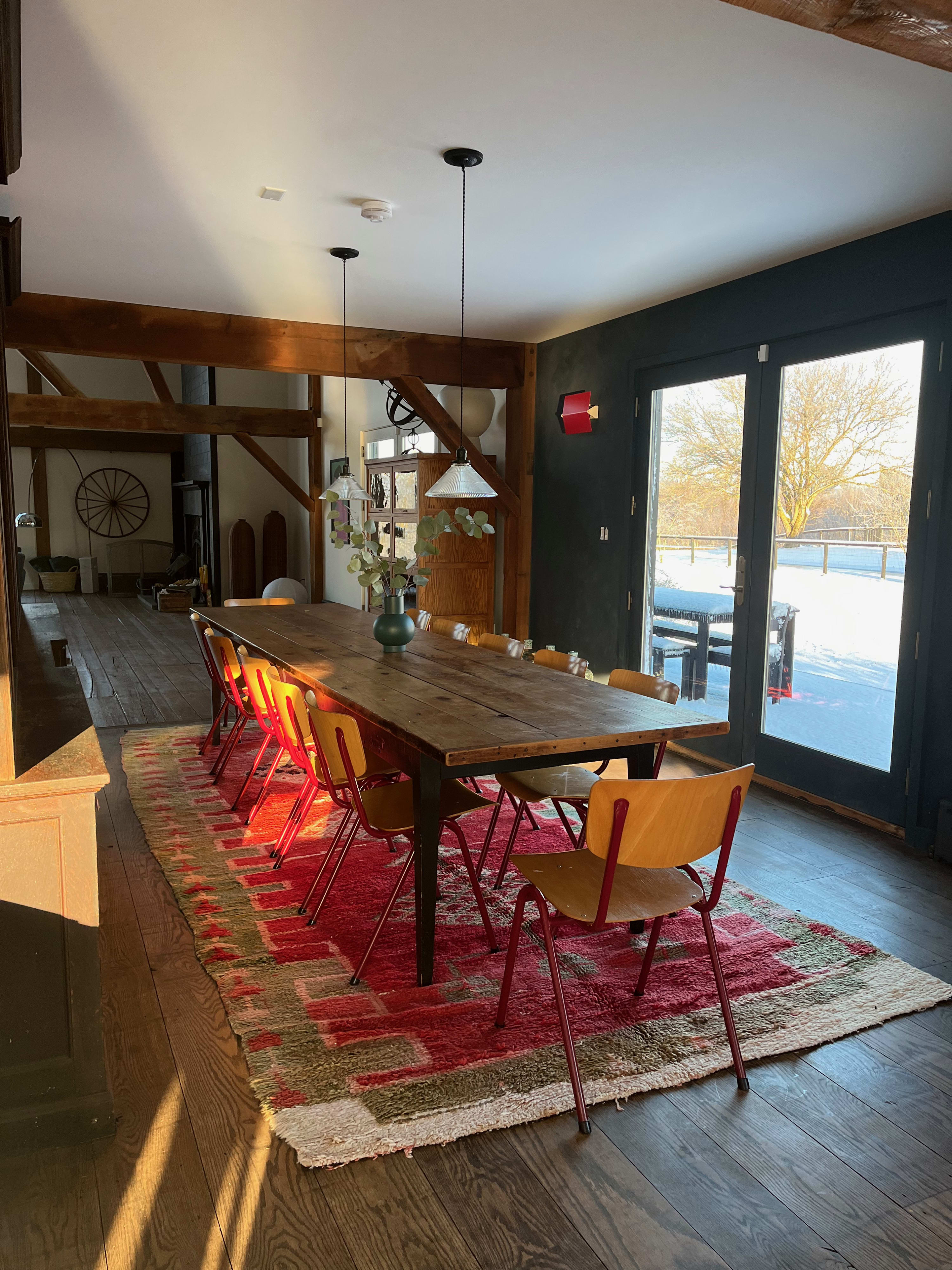 A sunlit dining room features a long, rustic wooden table with a vase of greenery, surrounded by bright orange and red chairs, all on a colorful patterned rug. Large glass doors open to a snowy outdoor landscape.