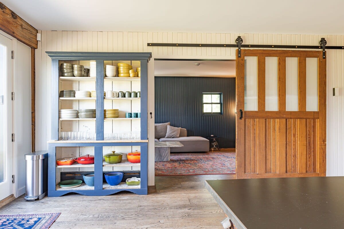 This view from the kitchen island shows a blue hutch filled with dishware, a sliding wooden barn door leading to another room, and light wood floors. The adjacent room features dark walls and a patterned rug.