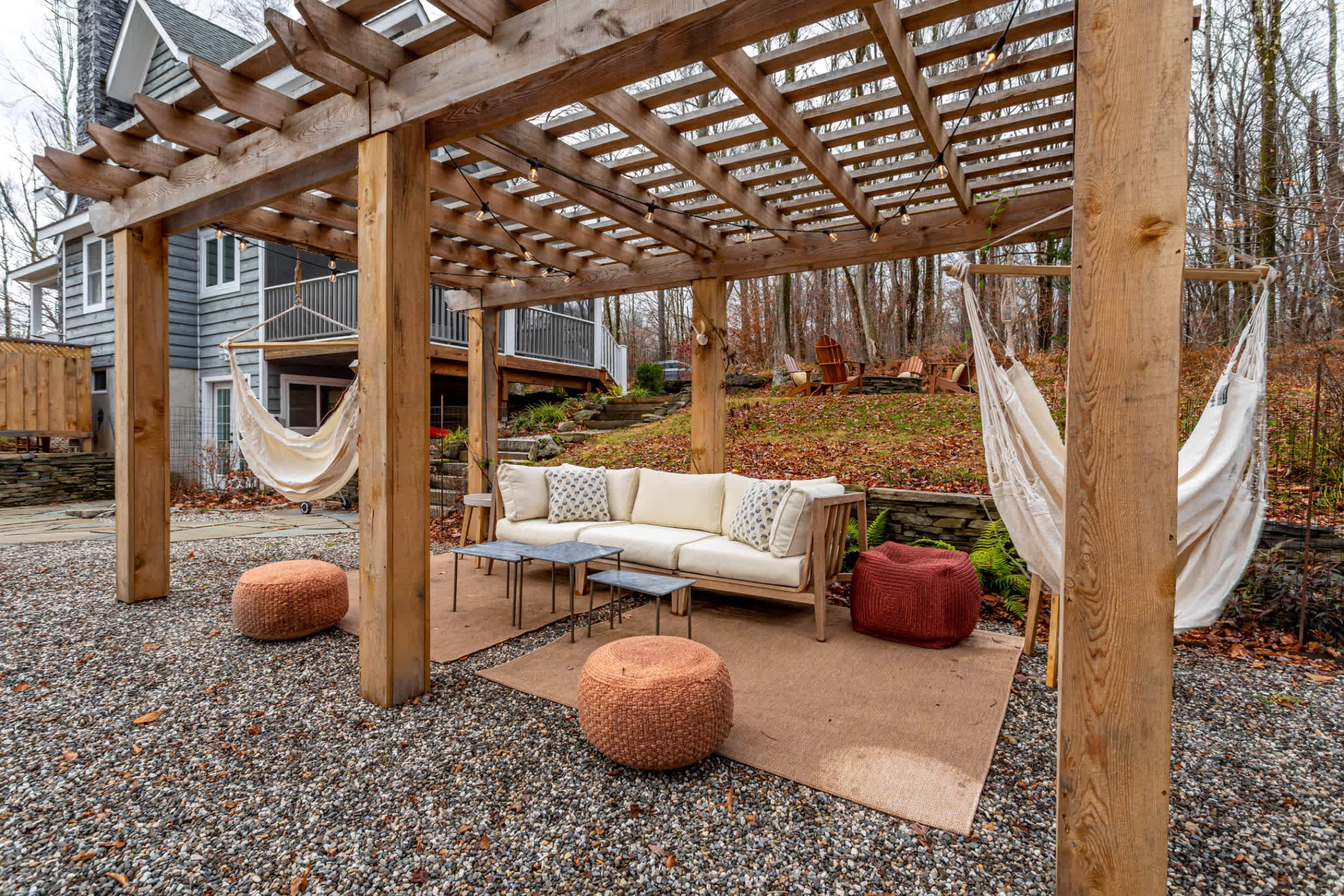 A spacious outdoor patio features a wooden pergola with string lights, providing shade over a comfortable outdoor sofa with cushions and two hammocks. The area is covered with gravel and includes decorative poufs, offering multiple relaxation spots.