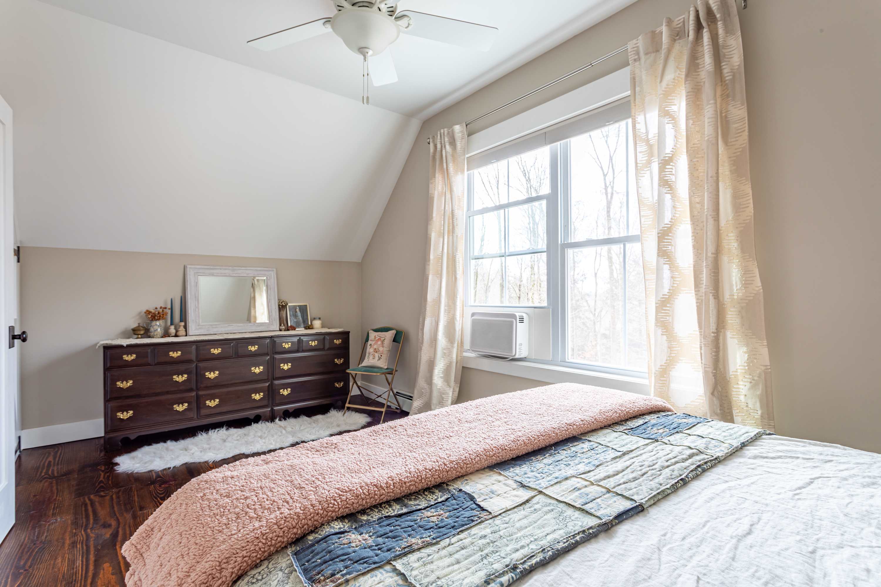 This bedroom showcases a dark wooden dresser with a mirror and decorative items, a large window with patterned curtains, and a ceiling fan, all set on dark hardwood floors under a sloped ceiling.
