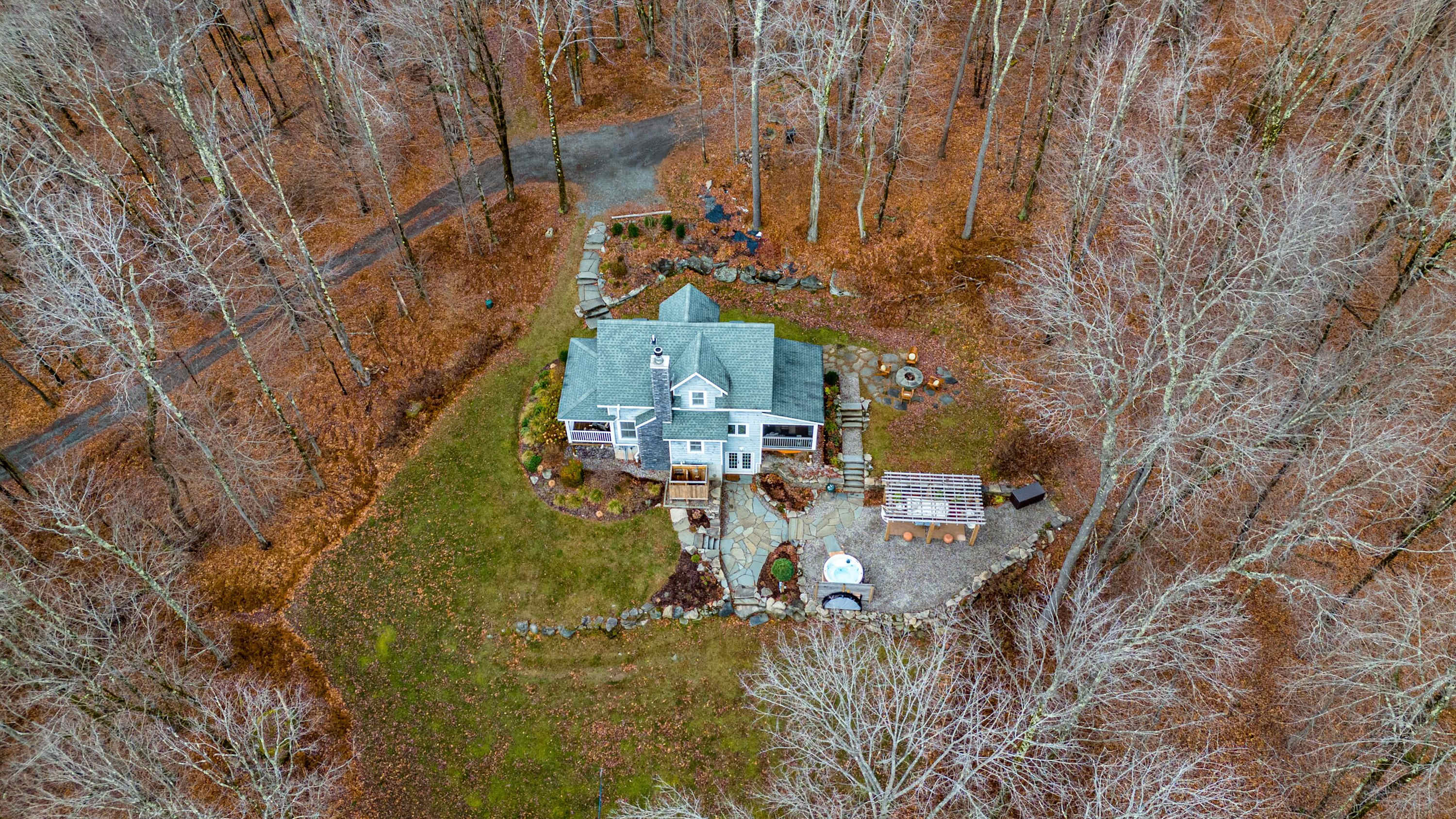 An aerial view showcases a gray-roofed house nestled in a dense, bare forest, surrounded by various outdoor amenities including a hot tub, fire pit, and pergola-covered seating areas, connected by paths.