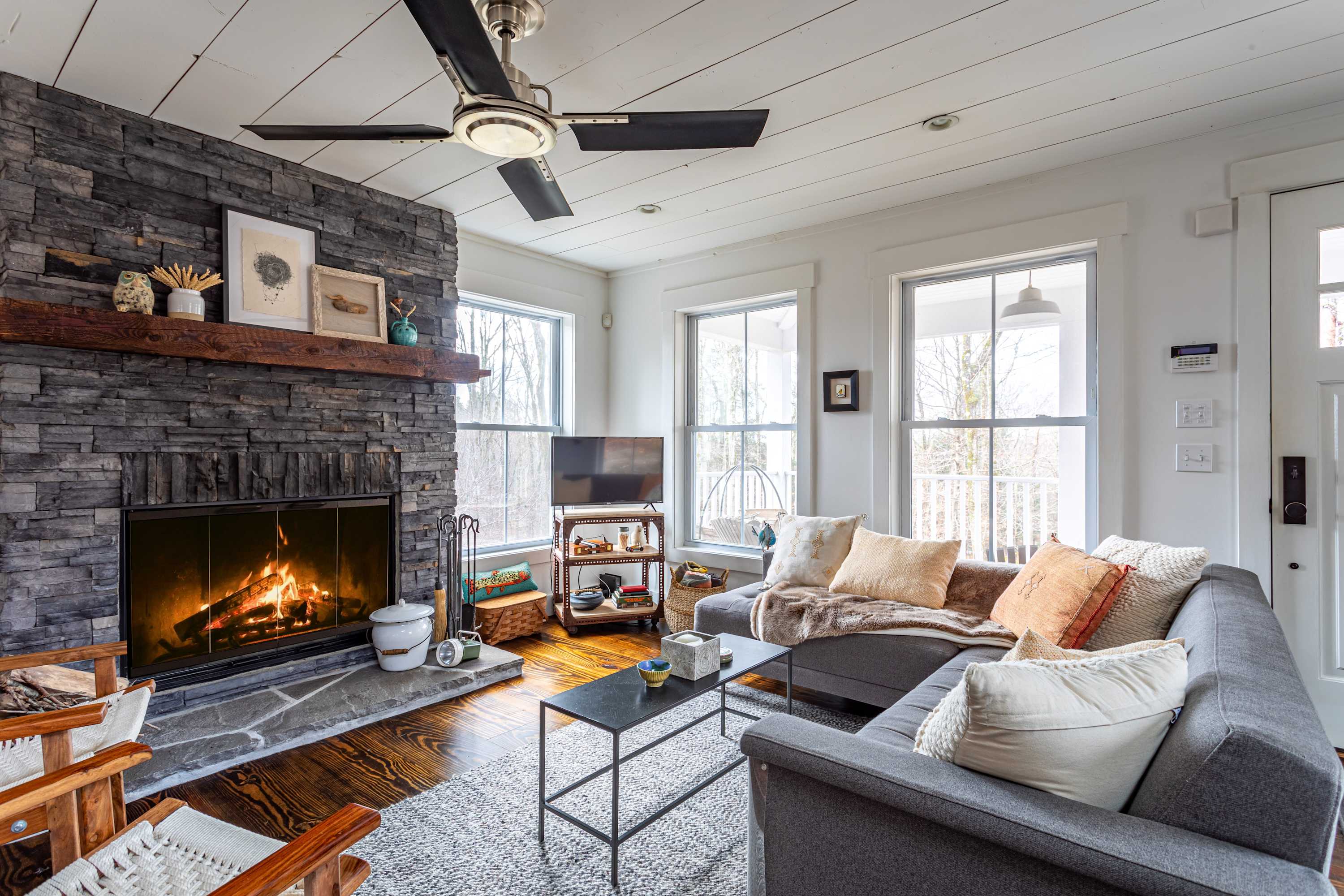Cozy living room with a large stone fireplace, dark wood floors, a grey sectional sofa with throw pillows, and a ceiling fan, illuminated by natural light from multiple windows.