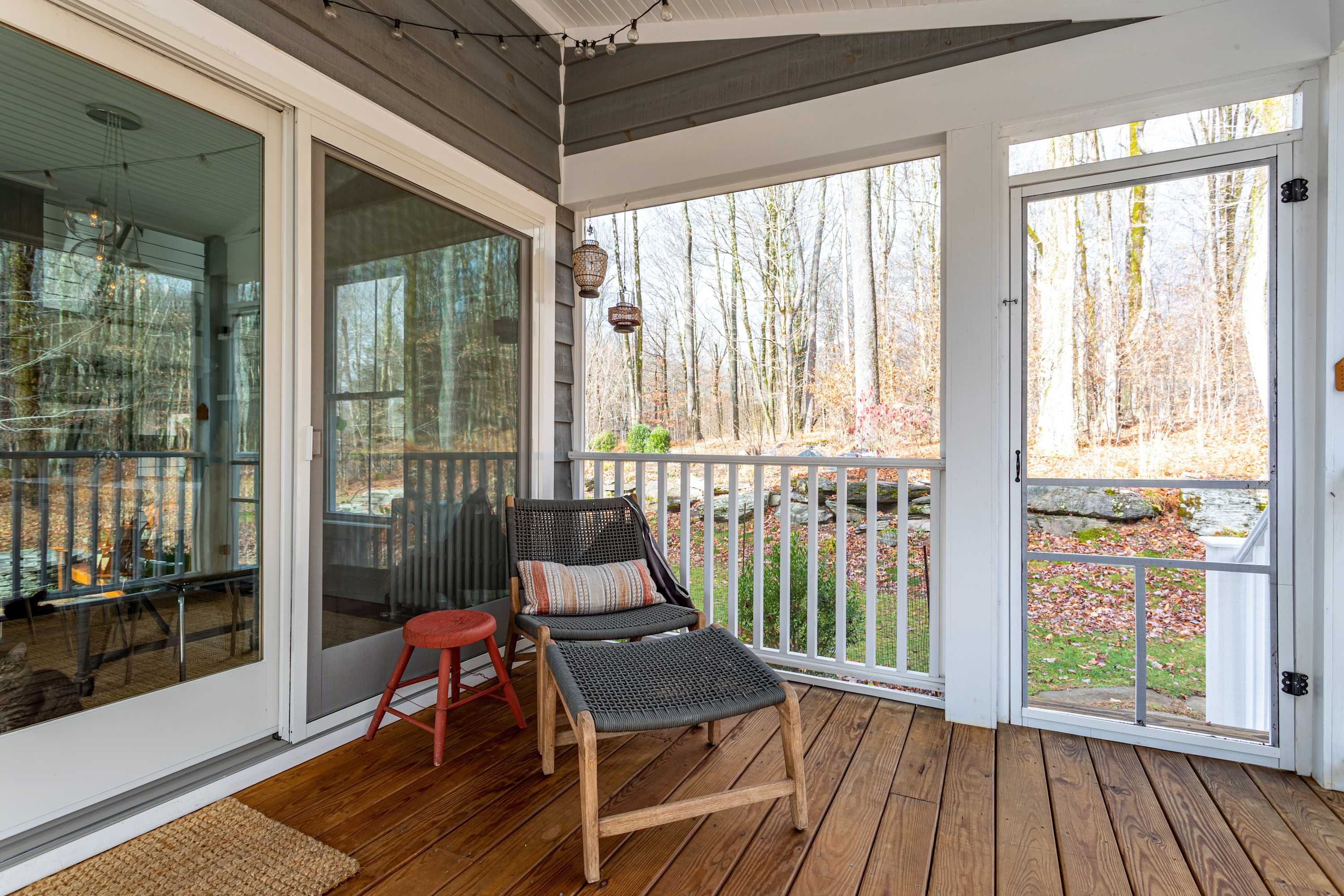A screened-in section of the porch features a modern woven lounge chair with a small red stool, offering a view of the natural, rocky backyard through the screen and railing.