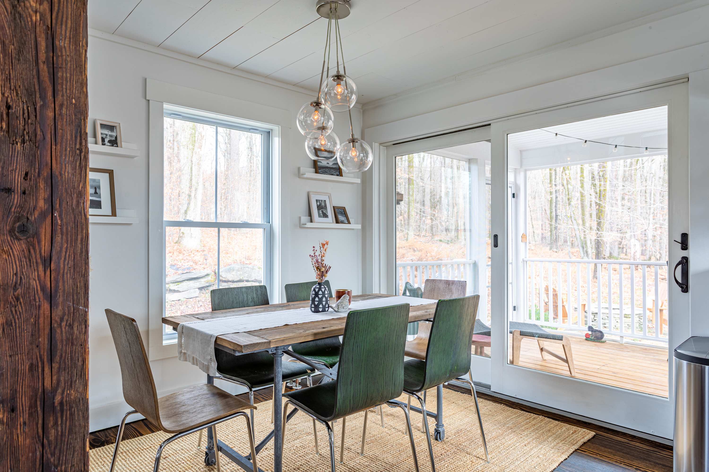 The dining area features a rustic wooden dining table with six chairs, set on a natural fiber rug, with a modern pendant light fixture overhead. Large windows and sliding glass doors lead to an outdoor deck, offering views of the surrounding woods.
