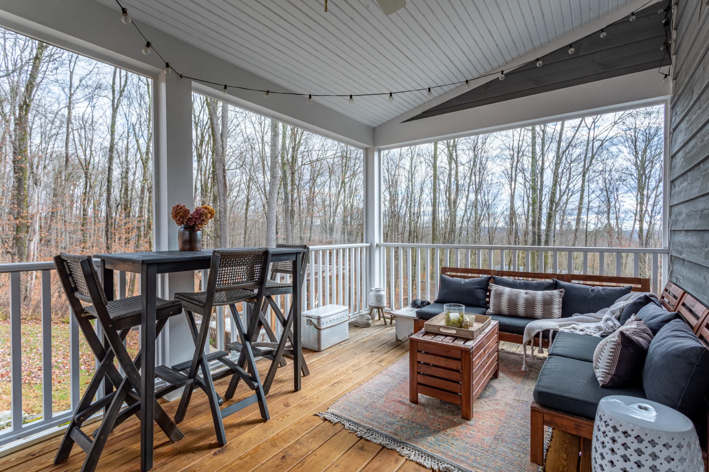 A spacious covered deck features a high-top bar table with stools and a comfortable outdoor sectional sofa with cushions, all set on warm wooden flooring. String lights add ambiance, and the deck overlooks a wooded area, providing a serene outdoor living space.